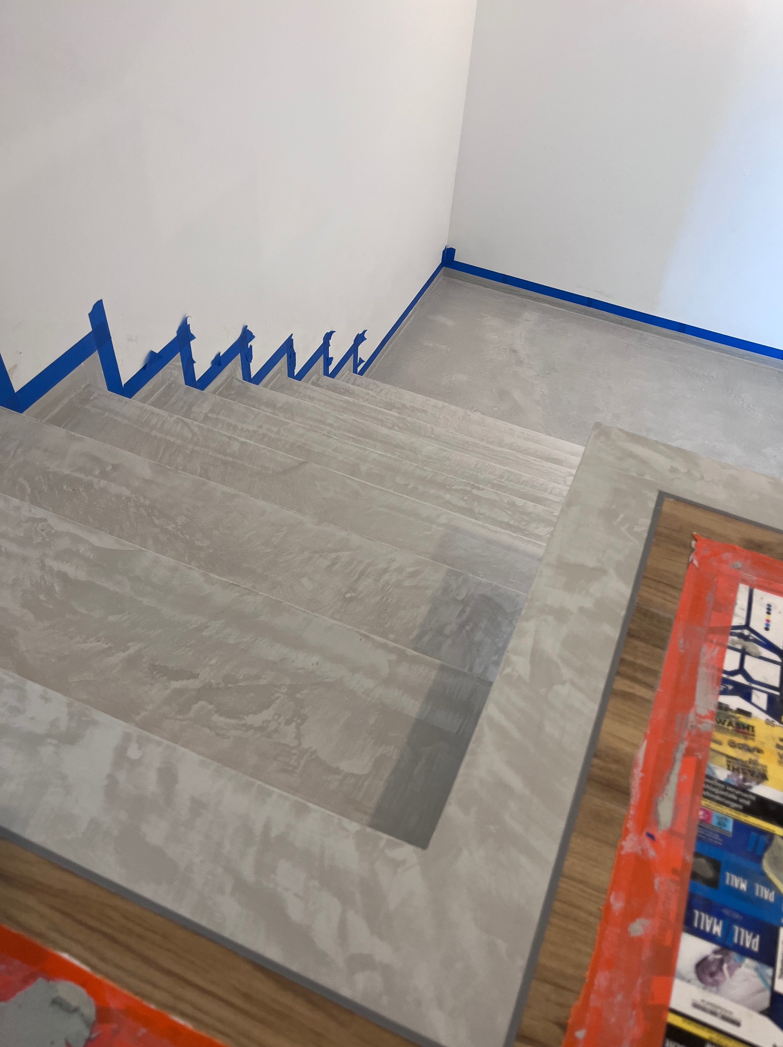Schody wykończone szarym mikrocementem, zabezpieczone niebieską taśmą malarską. Widok z góry, fragment podłogi z drewnopodobnych paneli.