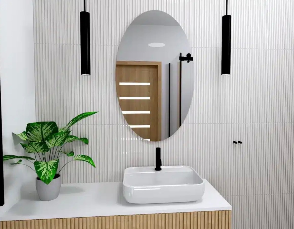 Owalne lustro z czarną armaturą nad białą umywalką w nowoczesnej łazience z białymi, żłobionymi płytkami i drewnianą szafką. Akcent stanowi zielona roślina doniczkowa.