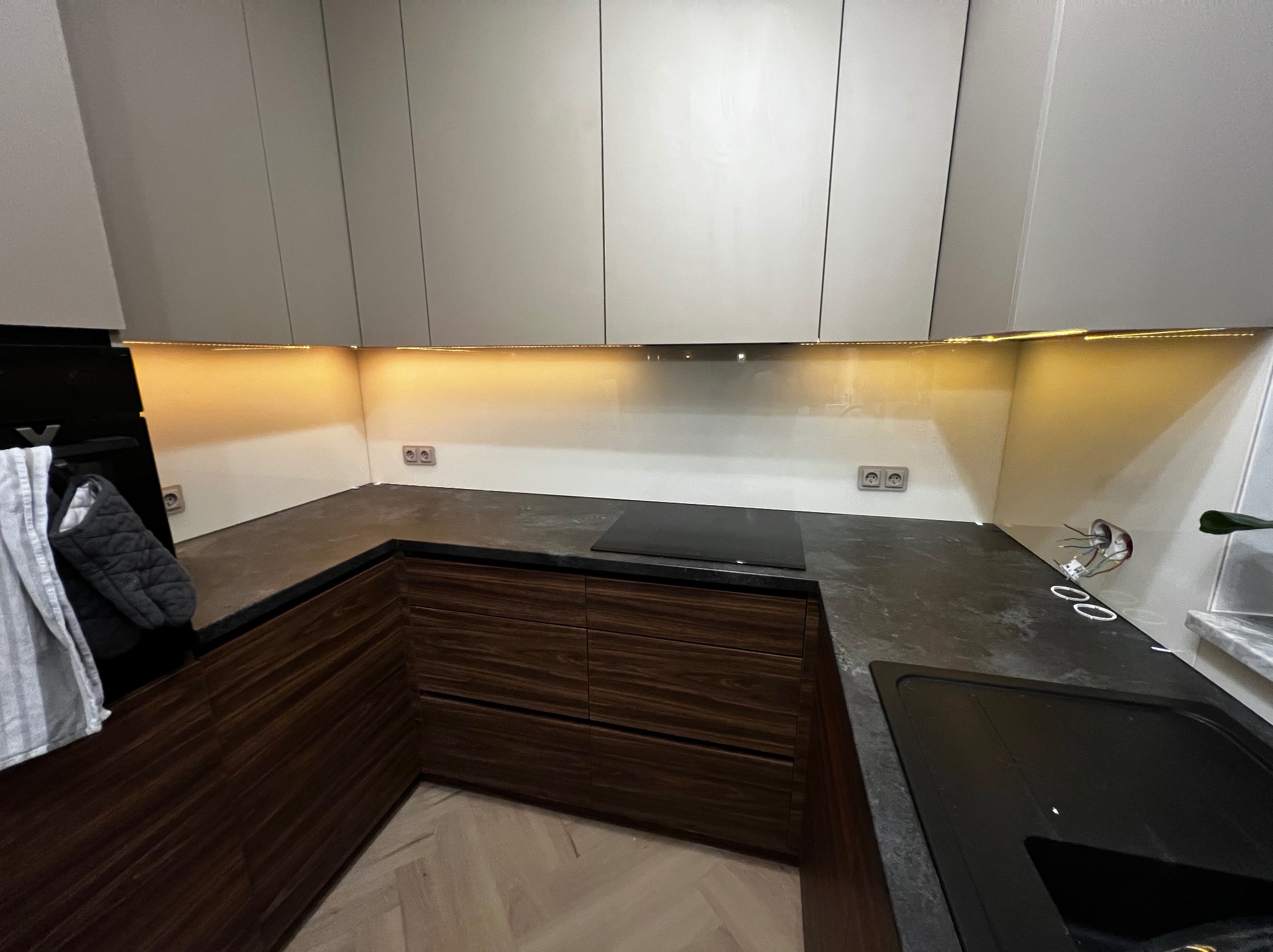 Nowoczesna kuchnia z szarymi szafkami i drewnianymi frontami, oświetlona listwą LED pod szafkami. Blat z ciemnego kamienia, widoczne gniazdka elektryczne.