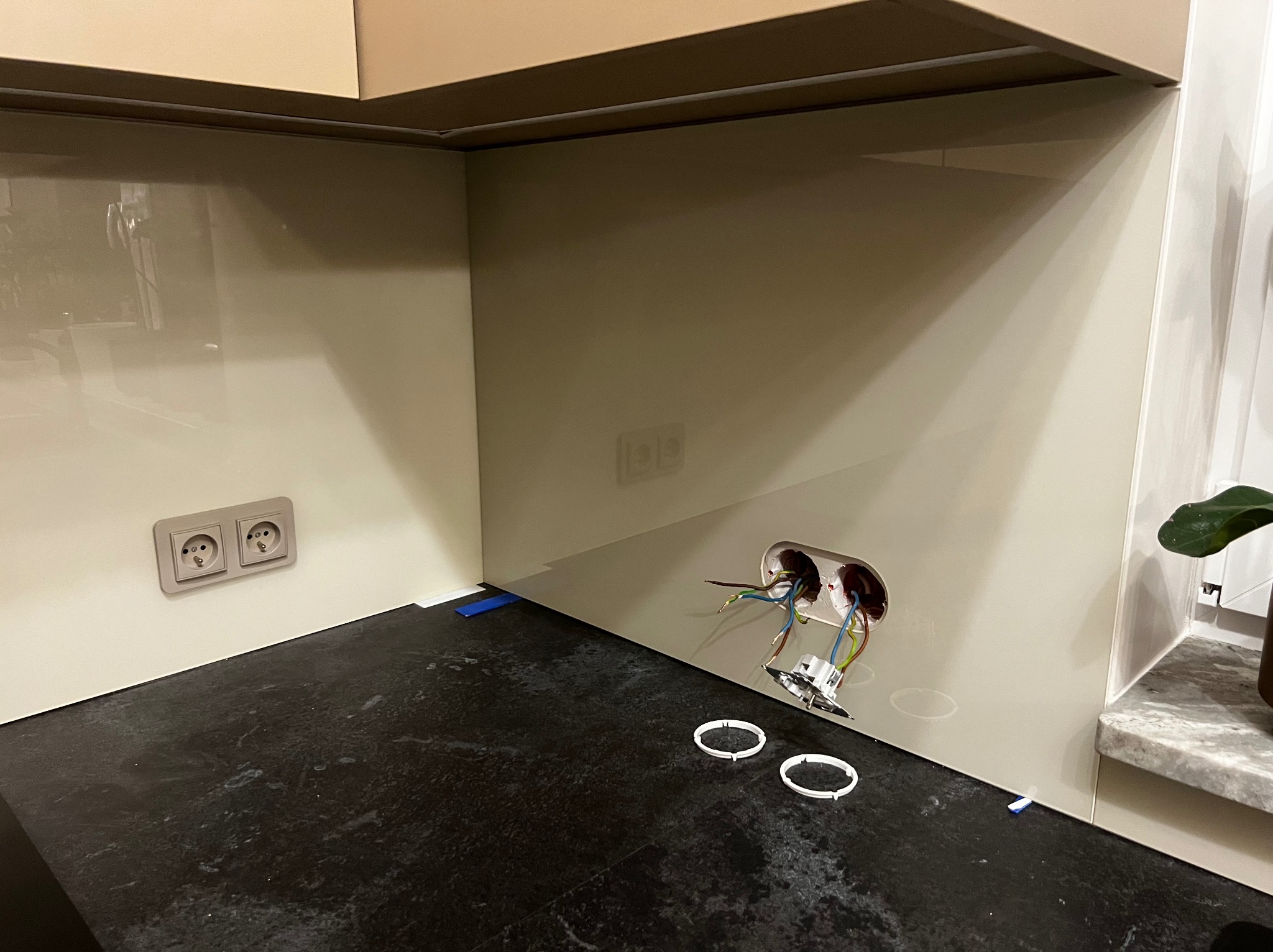 Montaż szklanej ściany w kuchni, widoczne gniazdka elektryczne i otwór na puszkę. Beżowe szkło, czarny blat. Etap prac wykończeniowych.