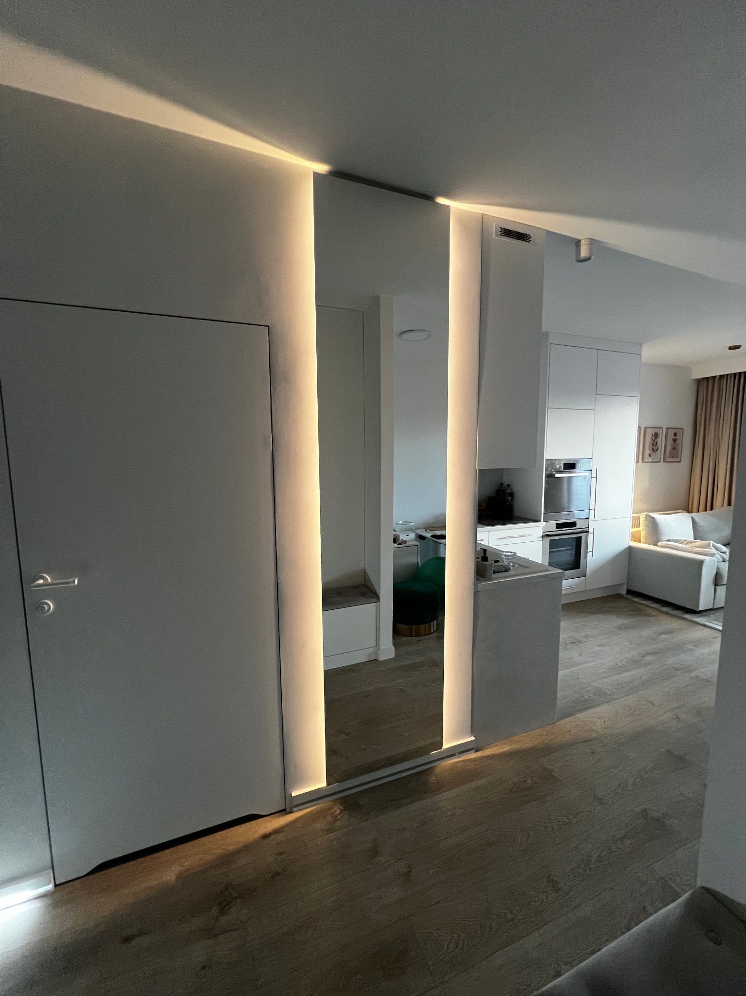 Nowoczesne lustro z podświetleniem LED w minimalistycznym wnętrzu. Widoczne odbicie kuchni i salonu, tworzące iluzję przestrzeni. Drzwi po lewej stronie kadru.