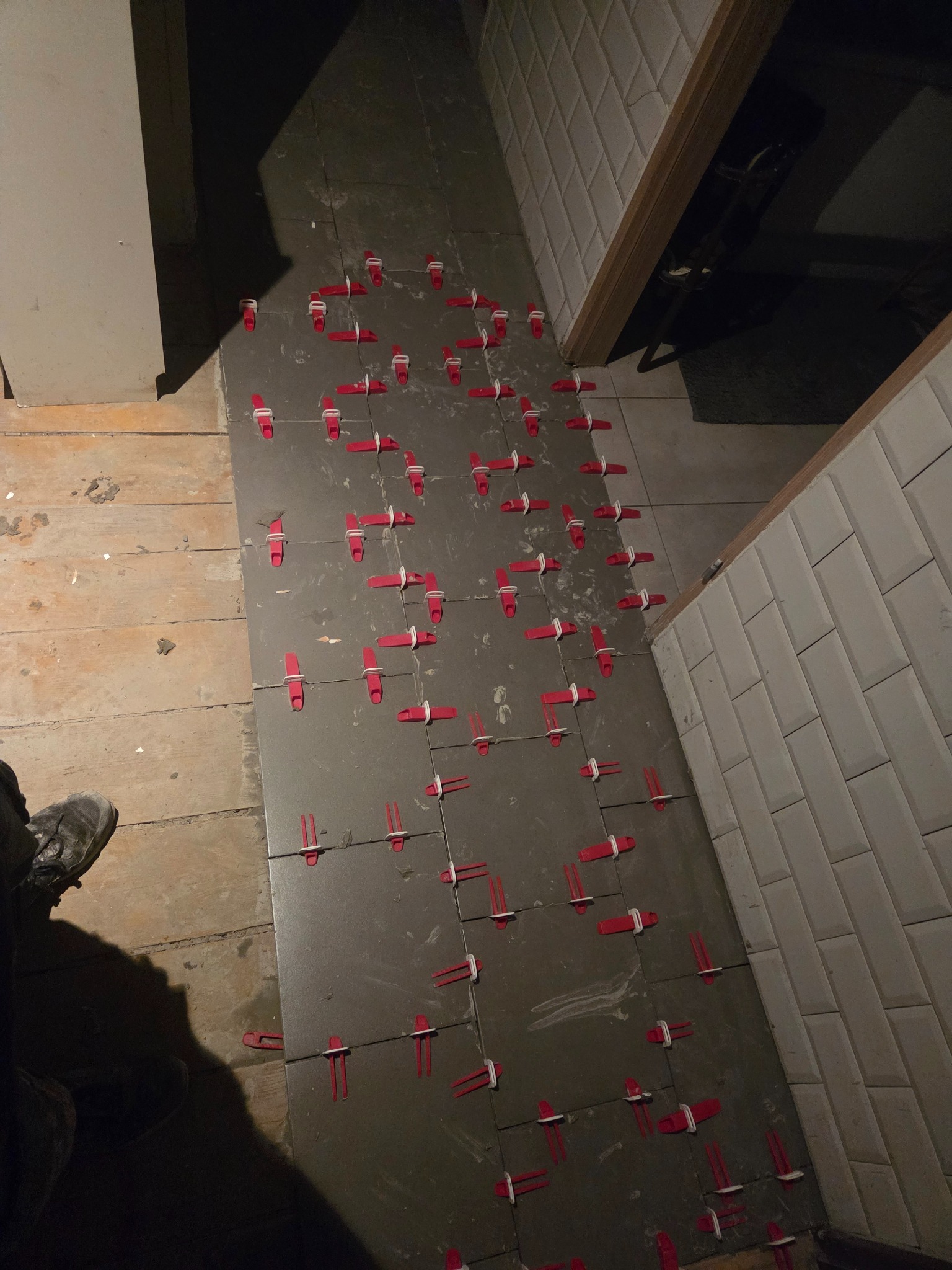 Układanie szarych płytek z użyciem czerwonych klipsów poziomujących. Widoczna część podłogi z desek. Fragment ściany wyłożonej białymi płytkami.