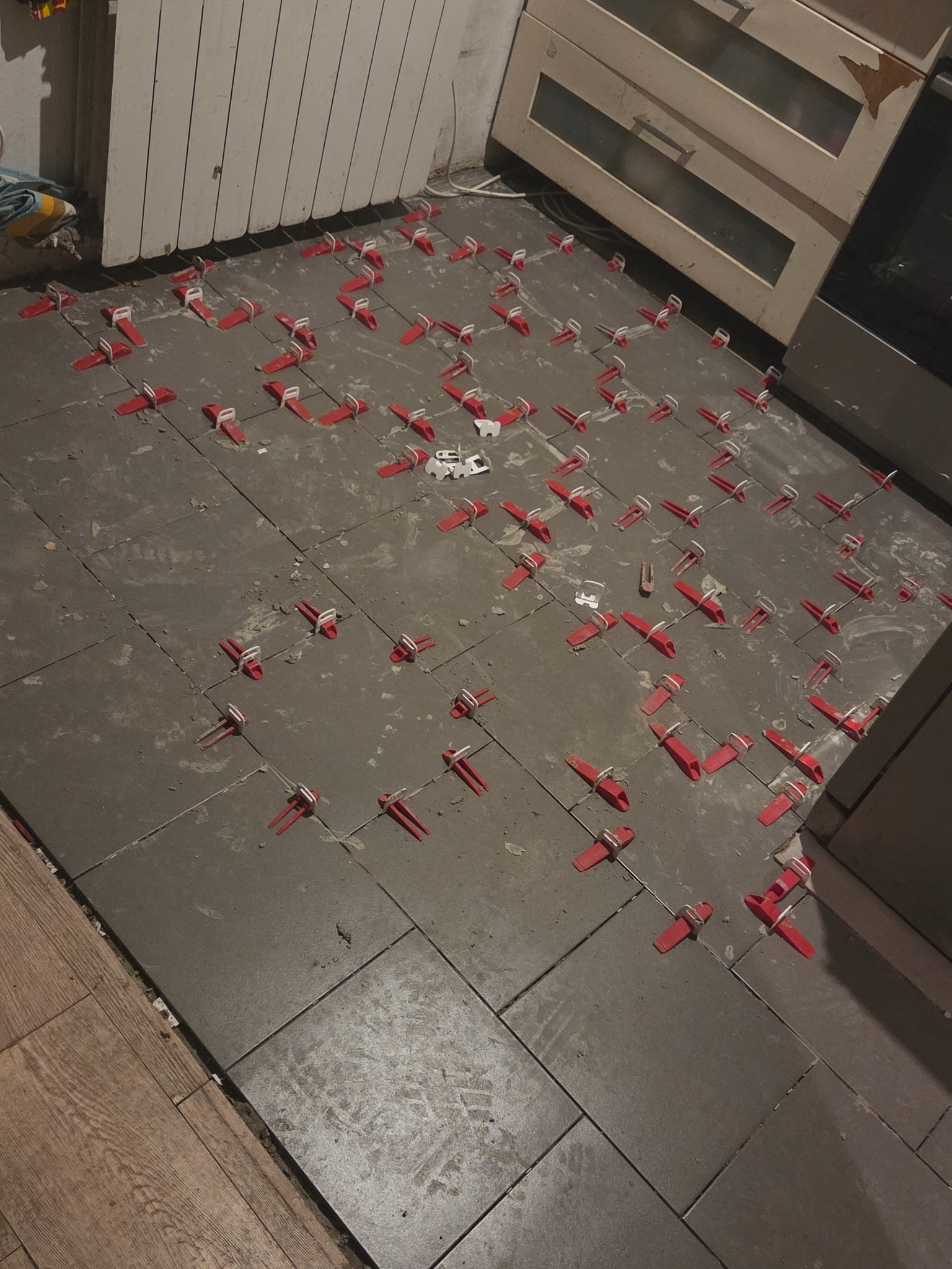 Szare płytki podłogowe w trakcie układania, z czerwonymi klipsami poziomującymi. Widoczne fragmenty drewnianej podłogi i mebli kuchennych. Ujęcie z góry, bliski kadr.