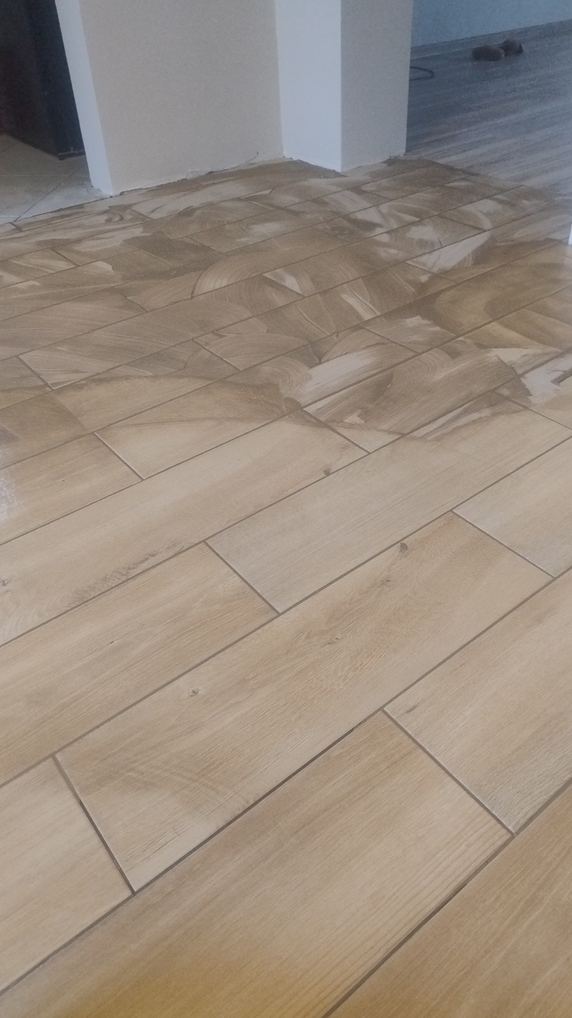 Ułożone, beżowe płytki podłogowe imitujące drewno, z widocznymi fugami i pozostałościami zaprawy, w jasnym wnętrzu. Ujęcie z niskiego kąta.