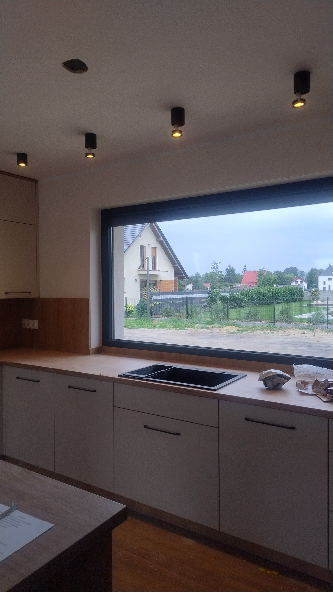 Nowoczesna kuchnia z oknem na ogród. Jasne szafki z czarnymi uchwytami, czarny zlew i punktowe oświetlenie sufitowe. Widoczny fragment niezakończonego montażu lampy.