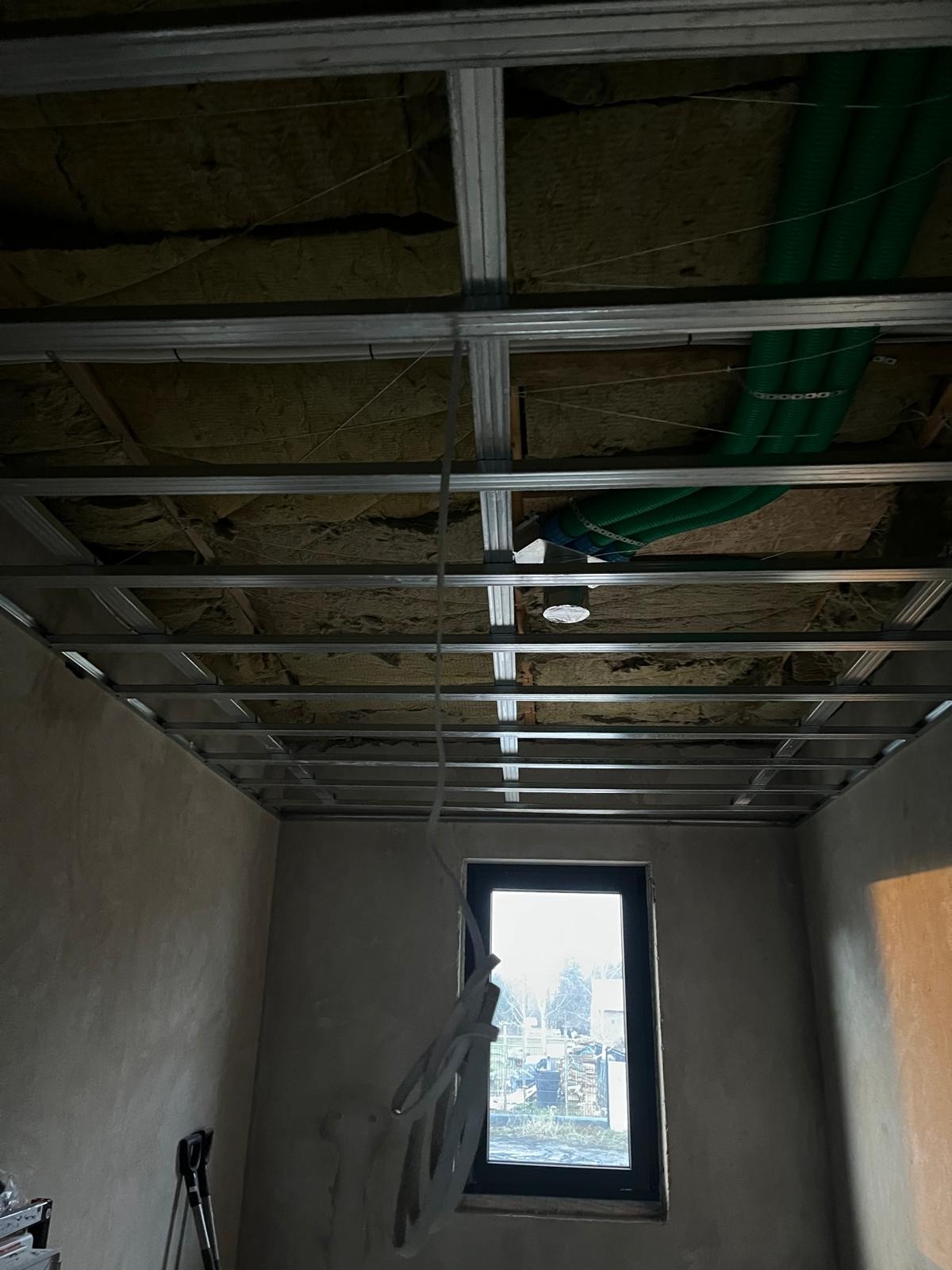 Sufit podwieszany z metalową konstrukcją, ociepleniem z wełny mineralnej i zielonymi rurami wentylacyjnymi, widok z dołu, surowe ściany, okno w tle.