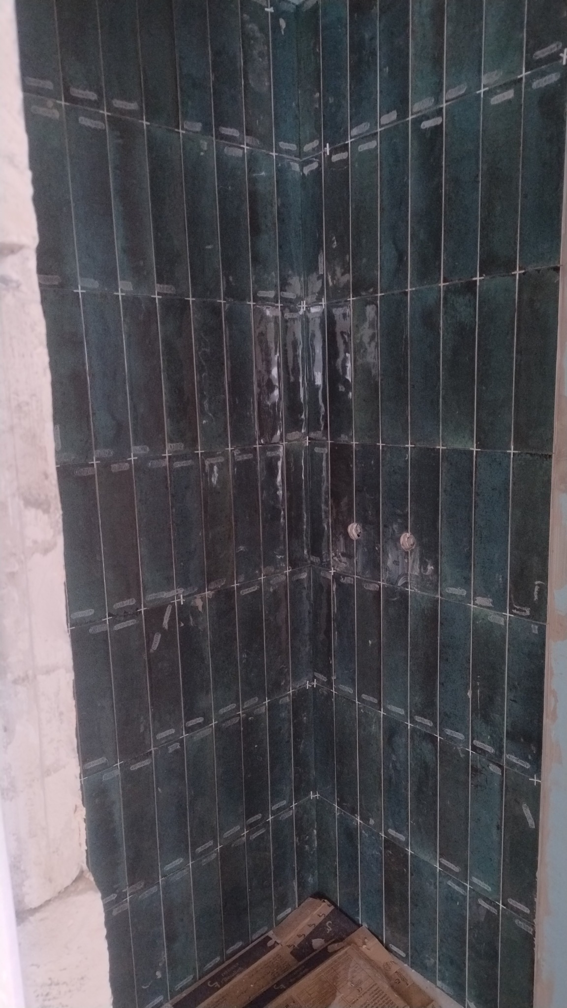 Ściana w trakcie układania ciemnozielonych, wąskich płytek w łazience. Widoczne krzyżyki dystansowe i fragment surowej ściany. Dwa otwory na instalacje.