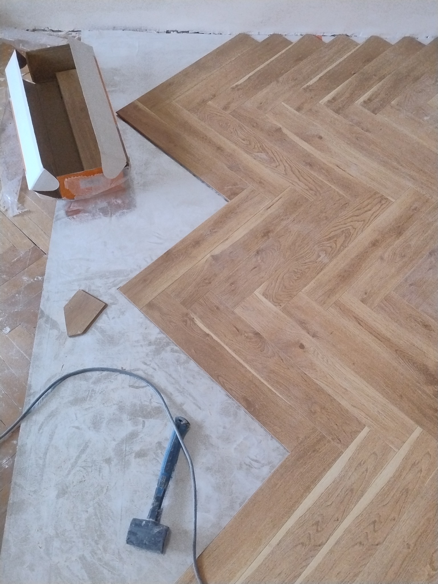 Układanie parkietu w jodełkę: fragment podłogi z ułożonym parkietem, obok karton, kabel i młotek gumowy. Widoczny etap prac wykończeniowych.