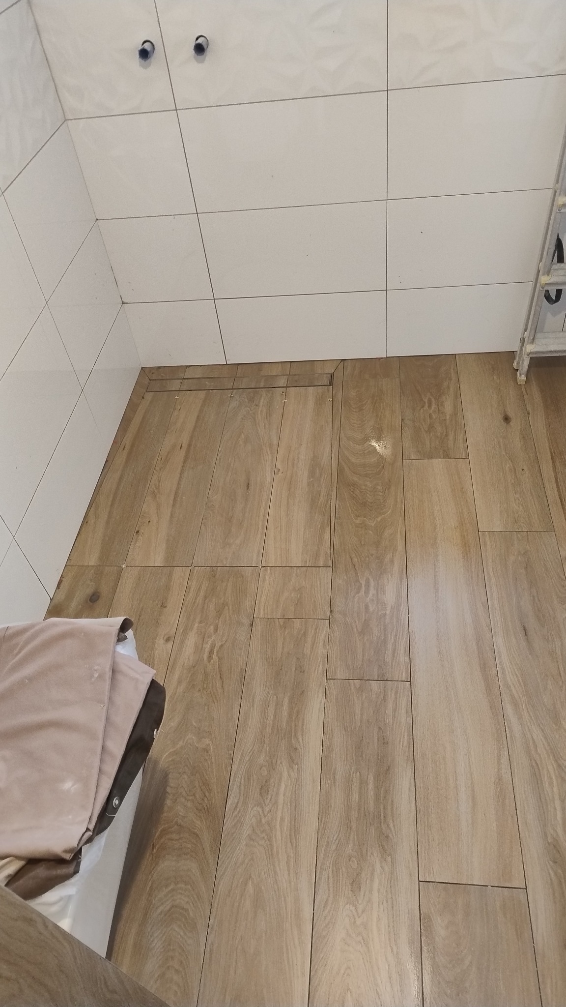 Ukończona podłoga z płytek imitujących drewno w łazience, białe płytki na ścianach, widoczne odpływy liniowe. Ujęcie z góry, fragment drabiny w rogu.