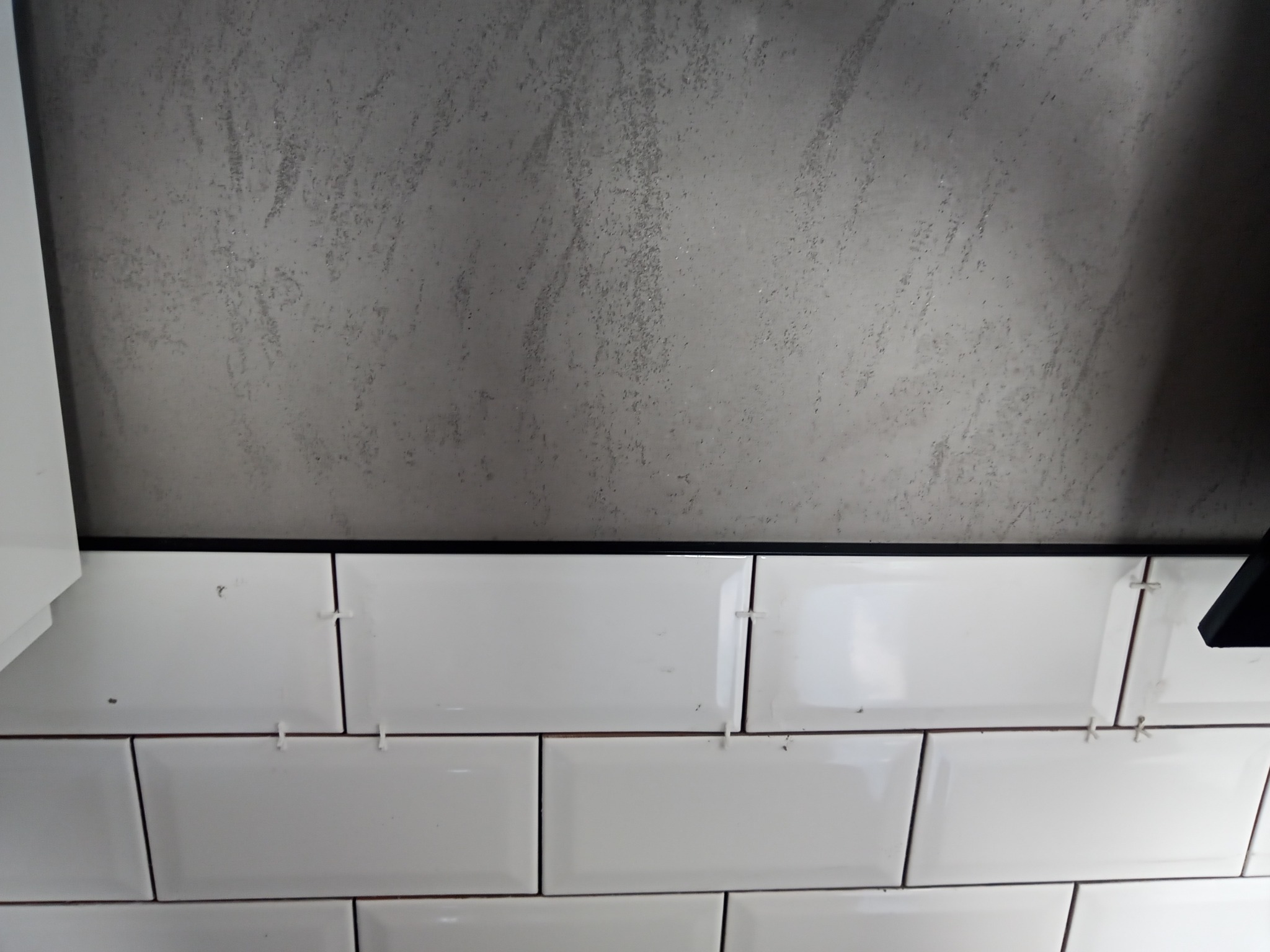 Białe, prostokątne płytki z czarną fugą, ułożone na ścianie pod szarą, teksturowaną powierzchnią. Widoczne krzyżyki dystansowe. Fragment białej szafki po lewej.