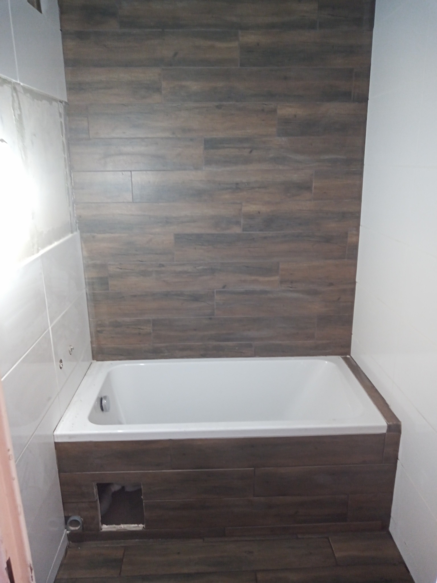 Biała wanna obudowana płytkami imitującymi drewno, z otworem rewizyjnym. Ściana wyłożona płytkami drewnopodobnymi do połowy wysokości, reszta biała.