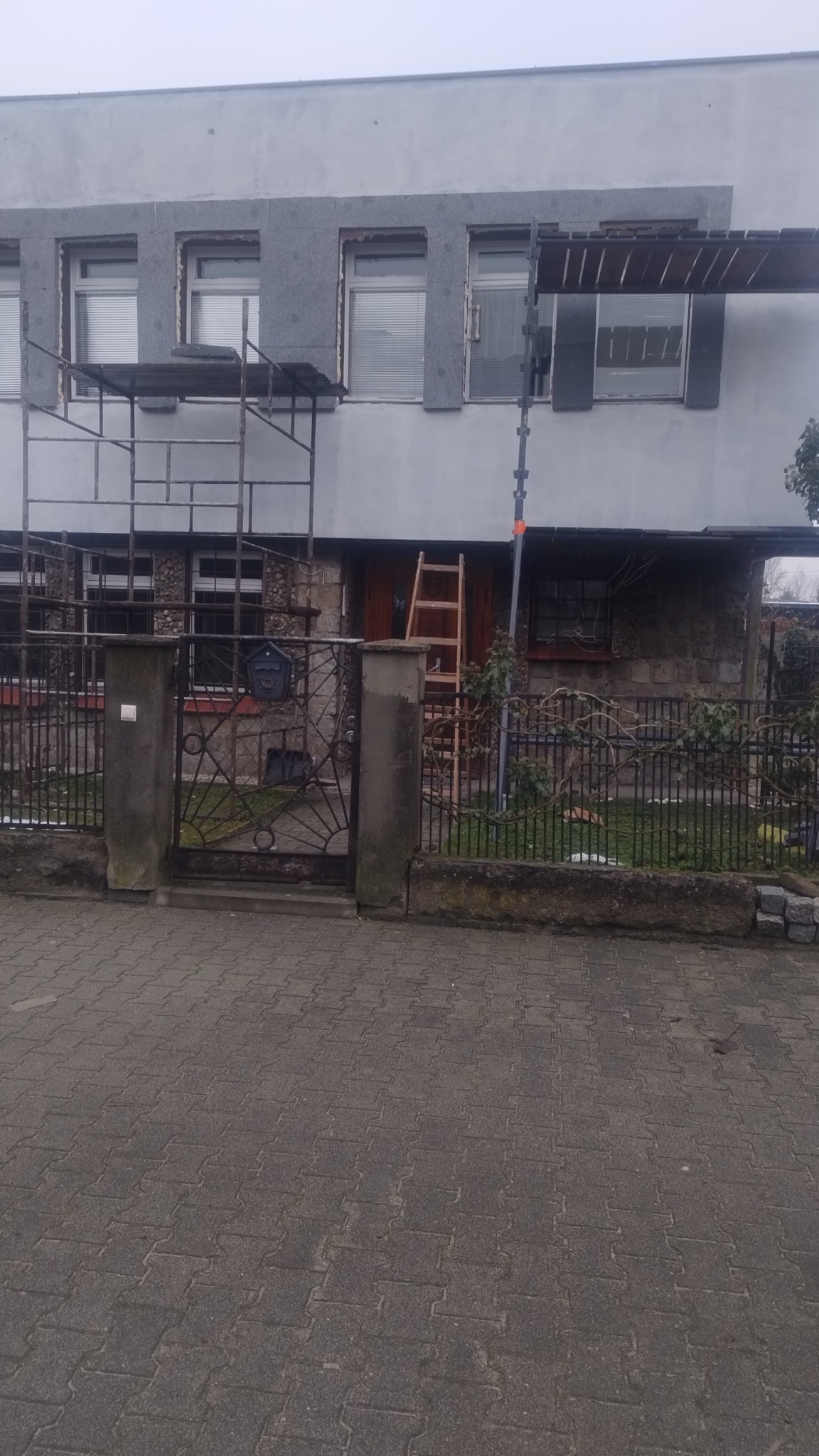 Ocieplanie elewacji budynku jednorodzinnego z użyciem rusztowania. Widoczne płyty izolacyjne wokół okien. Prace w toku, drabina oparta o ścianę.