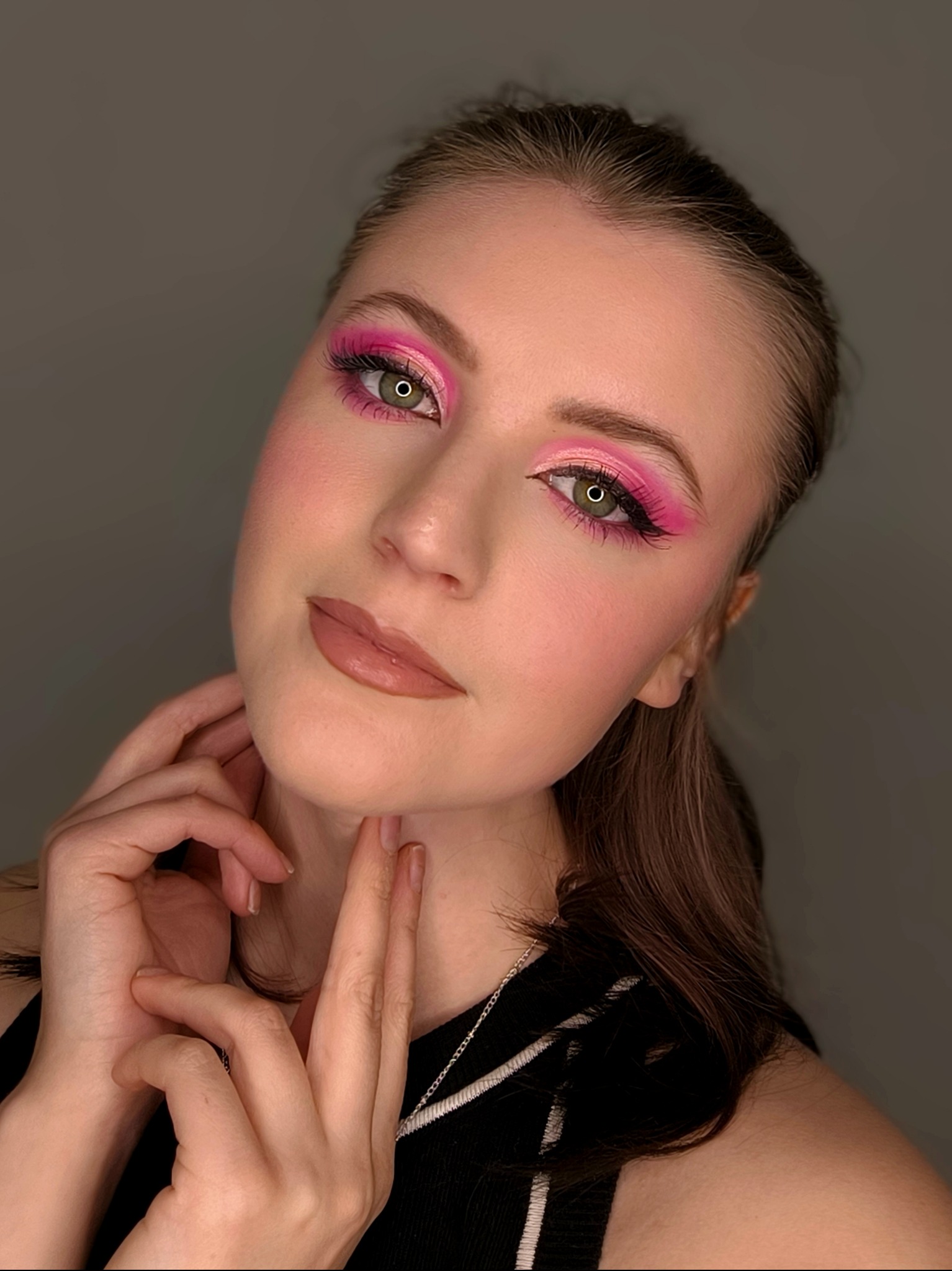 Portret kobiety z intensywnym, różowym makijażem oczu i błyszczącymi ustami. Dłonie delikatnie dotykają szyi, podkreślając stylizację. Subtelne oświetlenie.