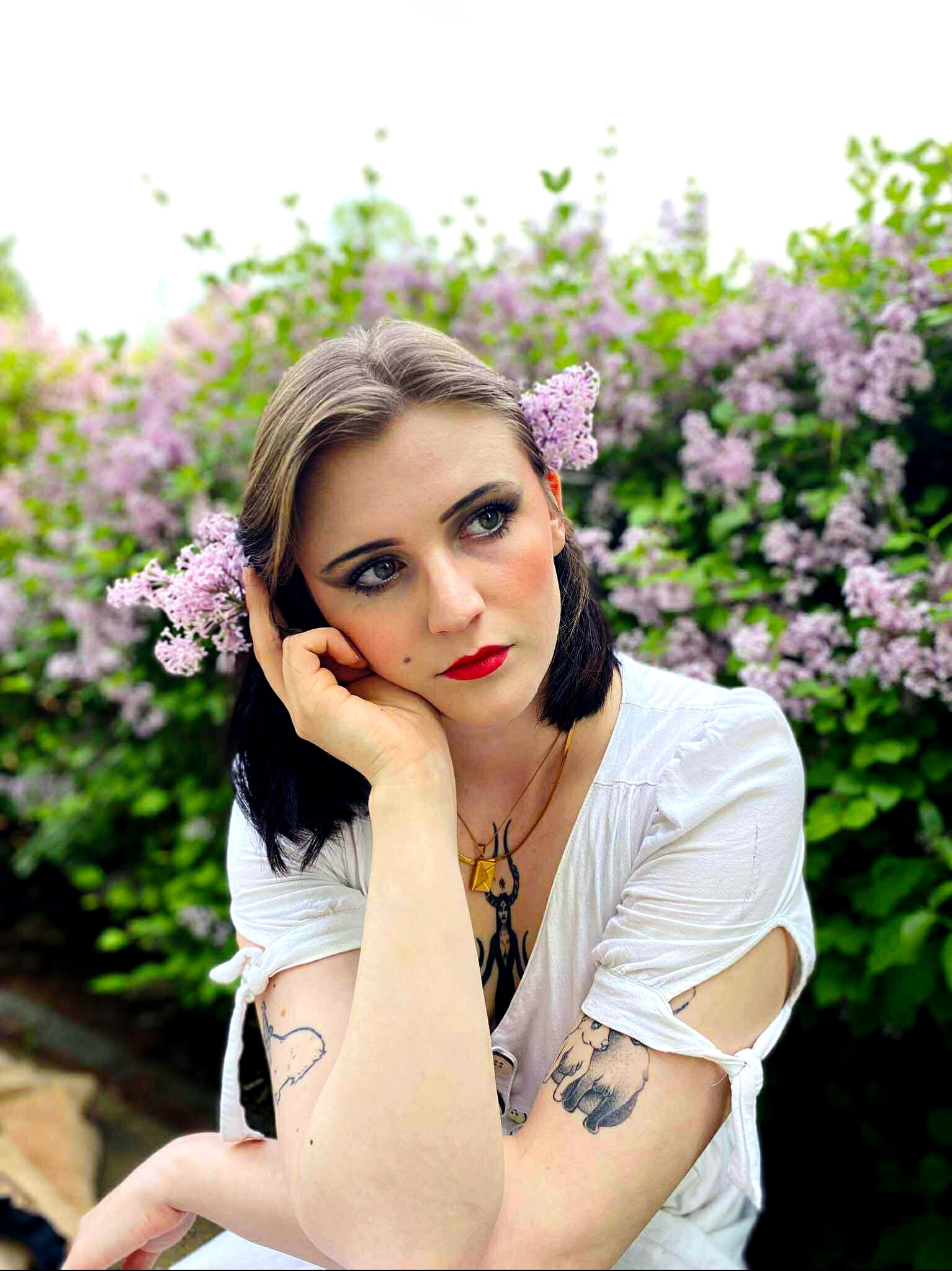 Portret kobiety z kwiatami bzu we włosach, z wyrazistym, czerwonym kolorem ust i delikatnym cieniem do powiek, pozującej na tle kwitnącego krzewu bzu.