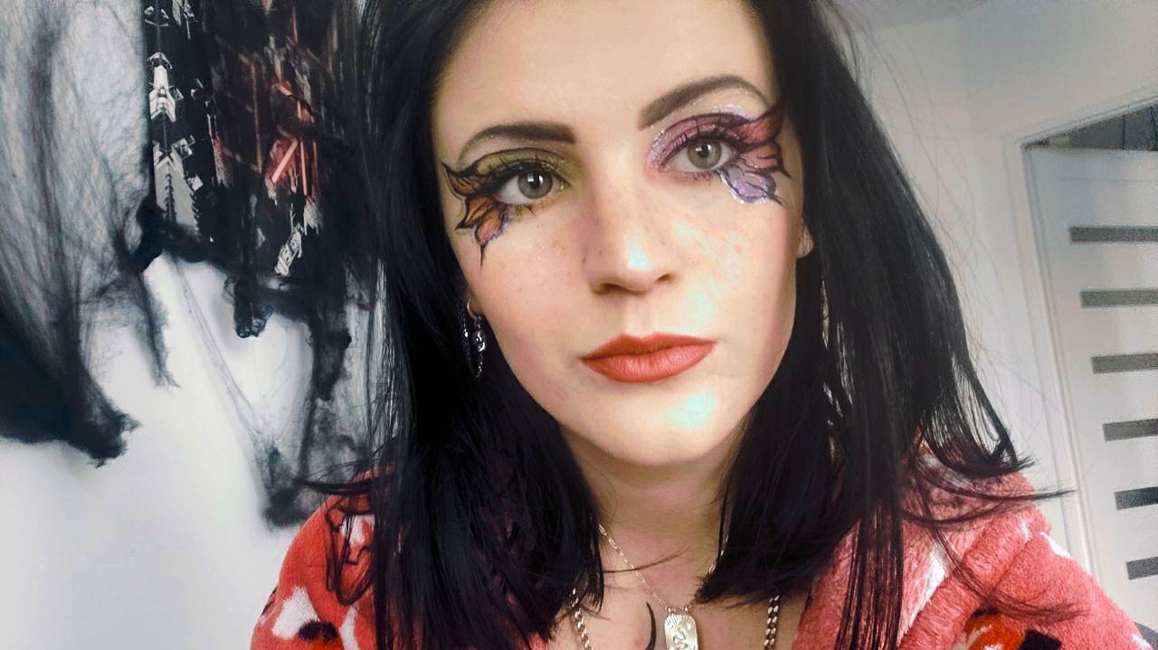 Portret kobiety z artystycznym, asymetrycznym zdobieniem oczu w stylu motyla, z czerwonymi ustami i czarnymi włosami, na tle dekoracji halloweenowych.