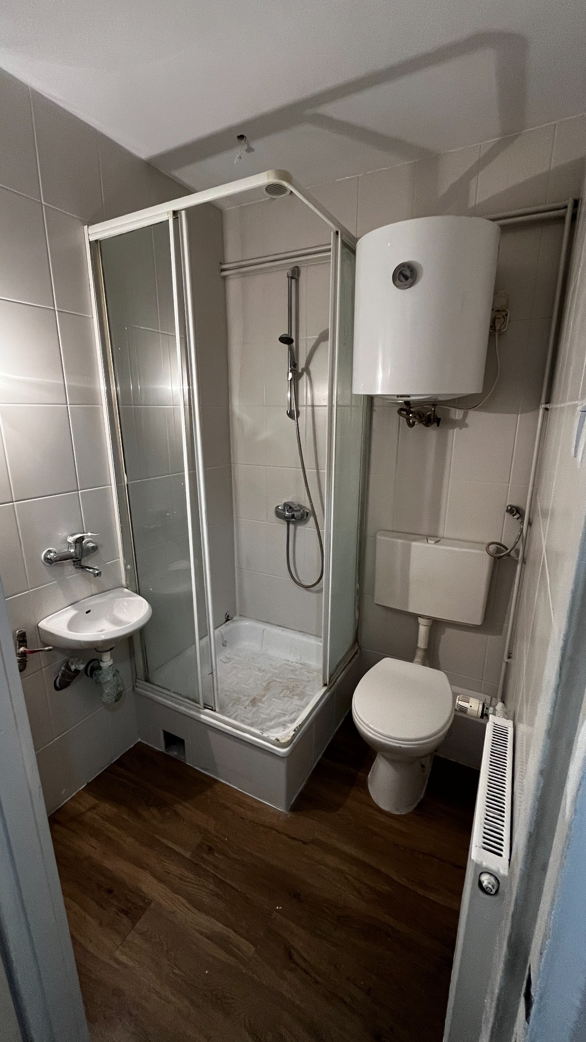 Wnętrze łazienki po remoncie: kabina prysznicowa, umywalka, toaleta i bojler. Ciemna podłoga kontrastuje z szarymi płytkami ściennymi. Widoczny grzejnik.