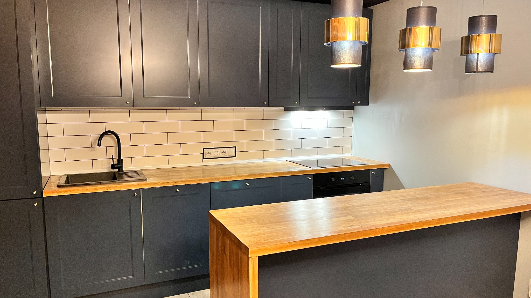 Nowoczesna kuchnia z szarymi szafkami, drewnianym blatem i białymi płytkami. Nad blatem wiszą trzy lampy z elementami w kolorze złota. Minimalistyczny design.