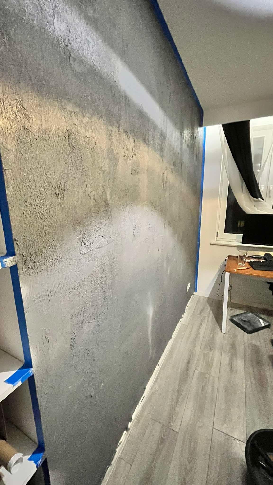 Ściana z efektem dekoracyjnym w szarościach, zabezpieczona niebieską taśmą malarską przy suficie i krawędziach. Widoczna podłoga z paneli i fragment biurka.