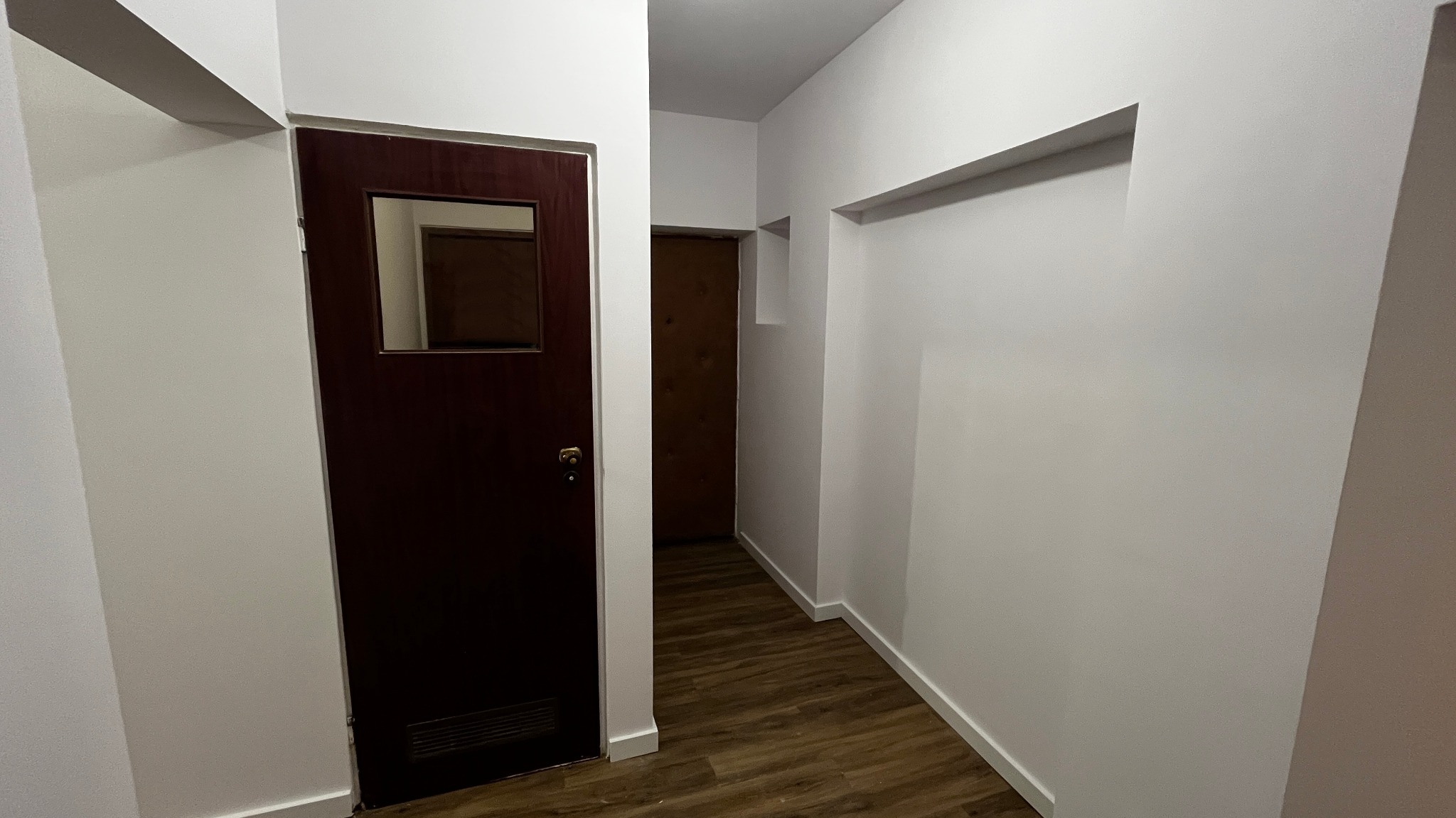 Korytarz po remoncie: białe ściany, drewnopodobna podłoga, ciemne drzwi z szybką. Widok na kolejne drzwi w głębi. Minimalistyczny design, świeżo po malowaniu.
