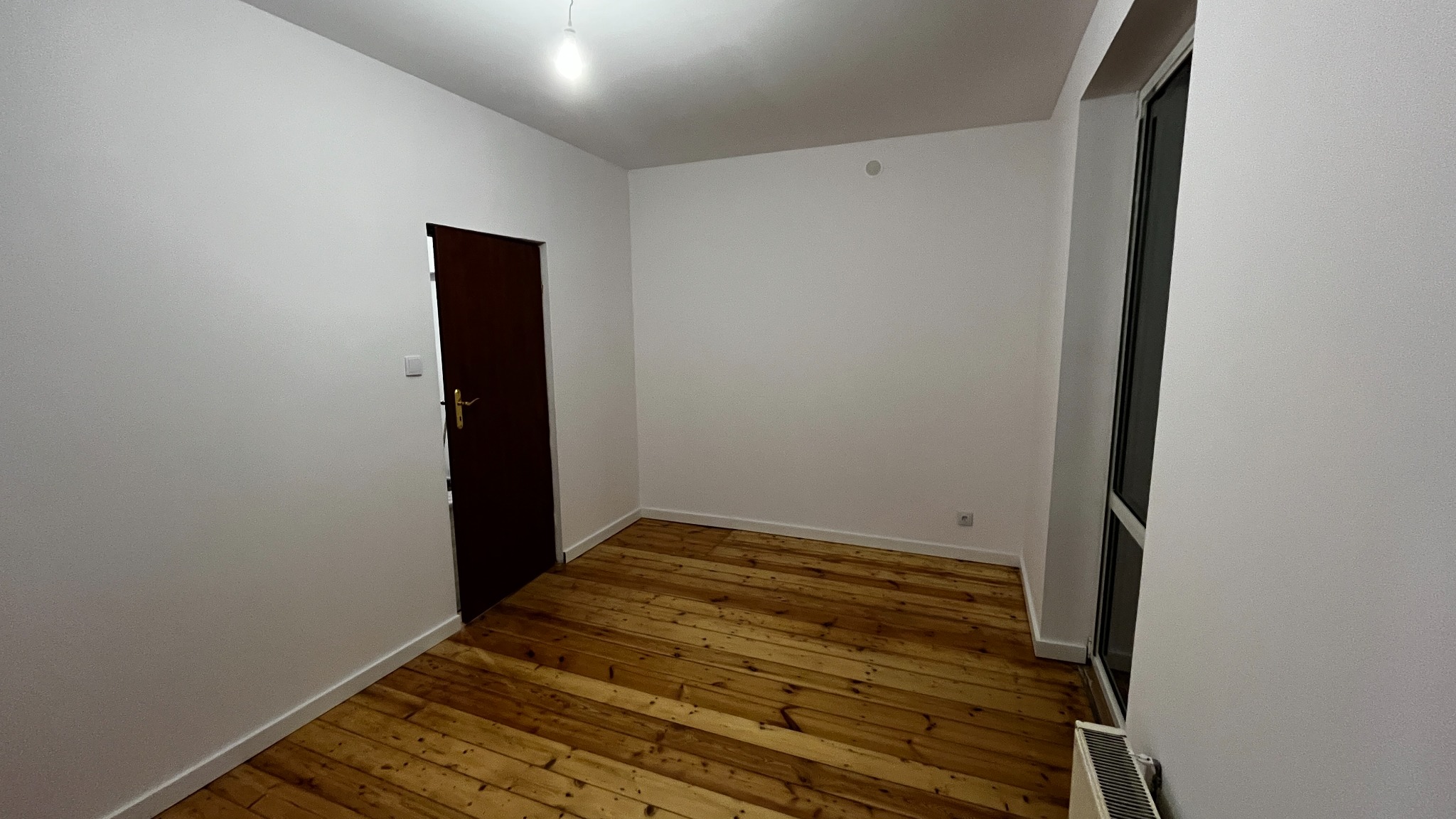 Puste, odnowione wnętrze z drewnianą podłogą, świeżo pomalowanymi ścianami i nowym oknem. Widoczna ciemna, zamknięta  furtka i  grzejnik.