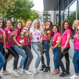Krełowski Foto-Art - Grupa kobiet w różowych koszulkach i jeansach pozuje z różami i rekwizytami na wieczór panieński. Panna młoda w welonie i koronie na tle nowoczesnego budynku.