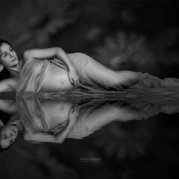Jacek Krełowski Photography - Czarno-białe, artystyczne zdjęcie kobiety w ciąży, leżącej na boku, owiniętej w delikatny materiał, z odbiciem w wodzie. Subtelne oświetlenie i kwiatowe tło dodają intymności.
