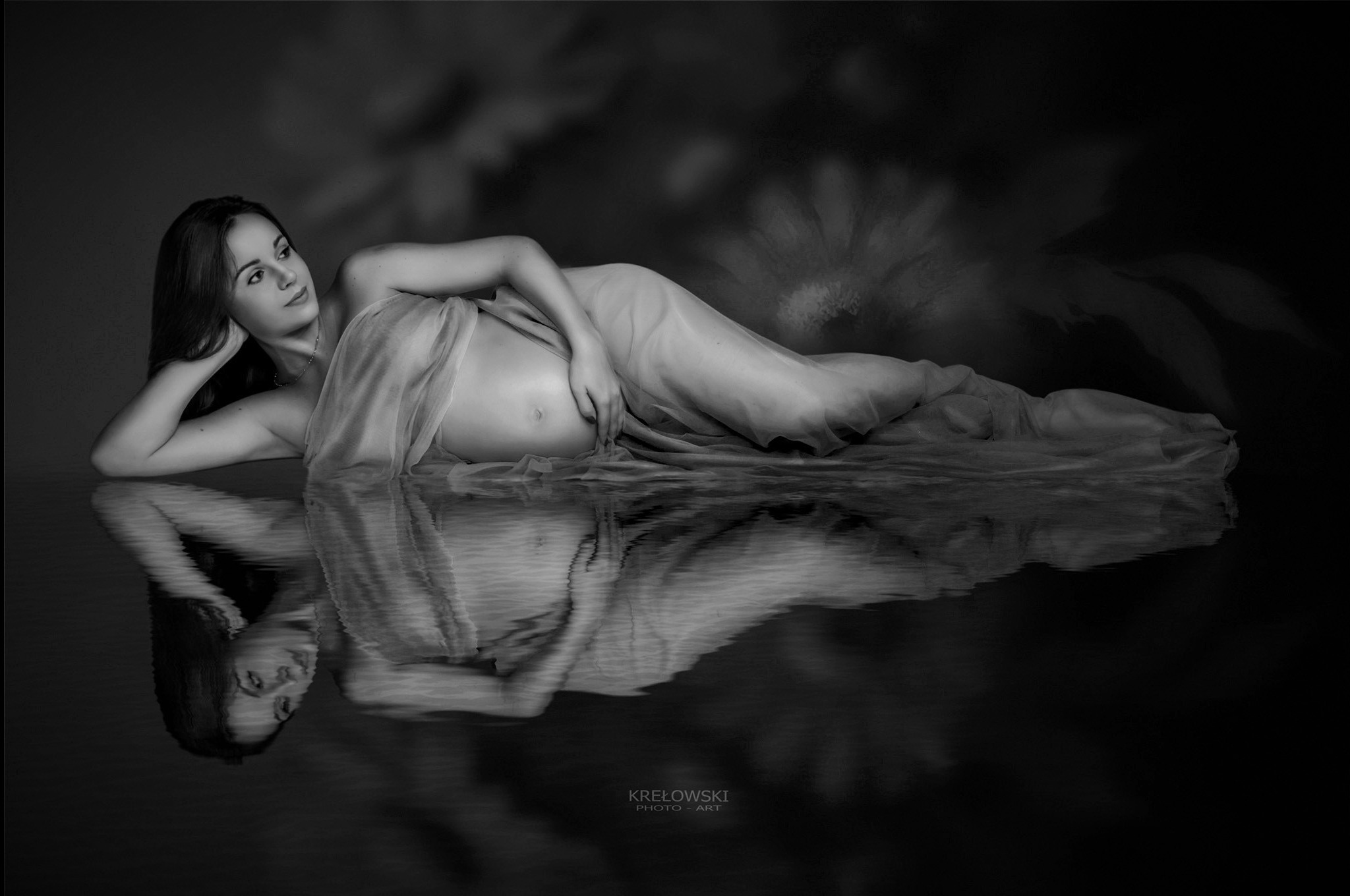 Czarno-białe, artystyczne zdjęcie kobiety w ciąży, leżącej na boku, owiniętej w delikatny materiał, z odbiciem w wodzie. Subtelne oświetlenie i kwiatowe tło dodają intymności.