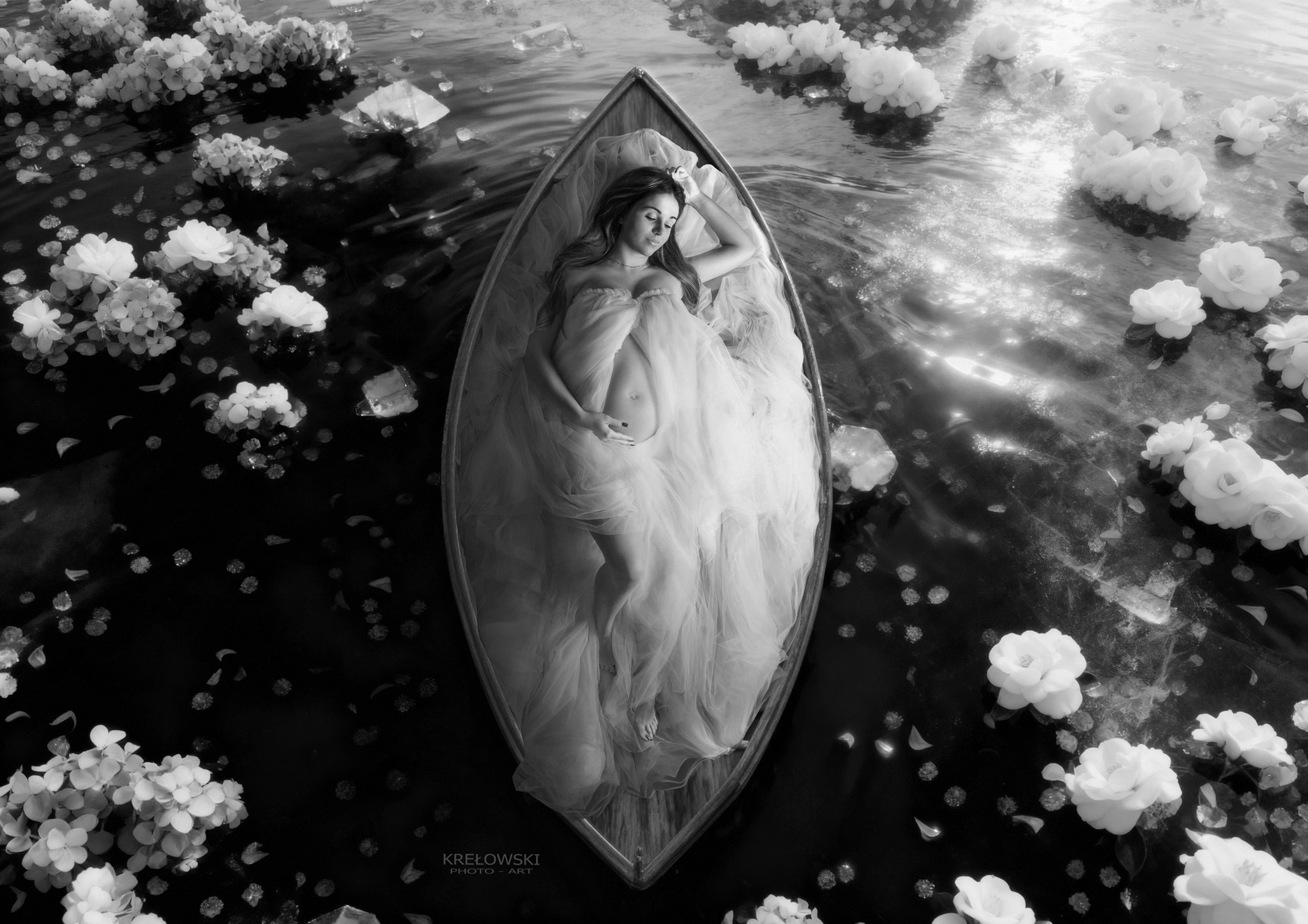 Czarno-białe, artystyczne ujęcie z góry: kobieta w ciąży, otulona woalem, leży w łodzi na wodzie, wokół pływają białe kwiaty. Delikatne światło podkreśla intymność chwili.