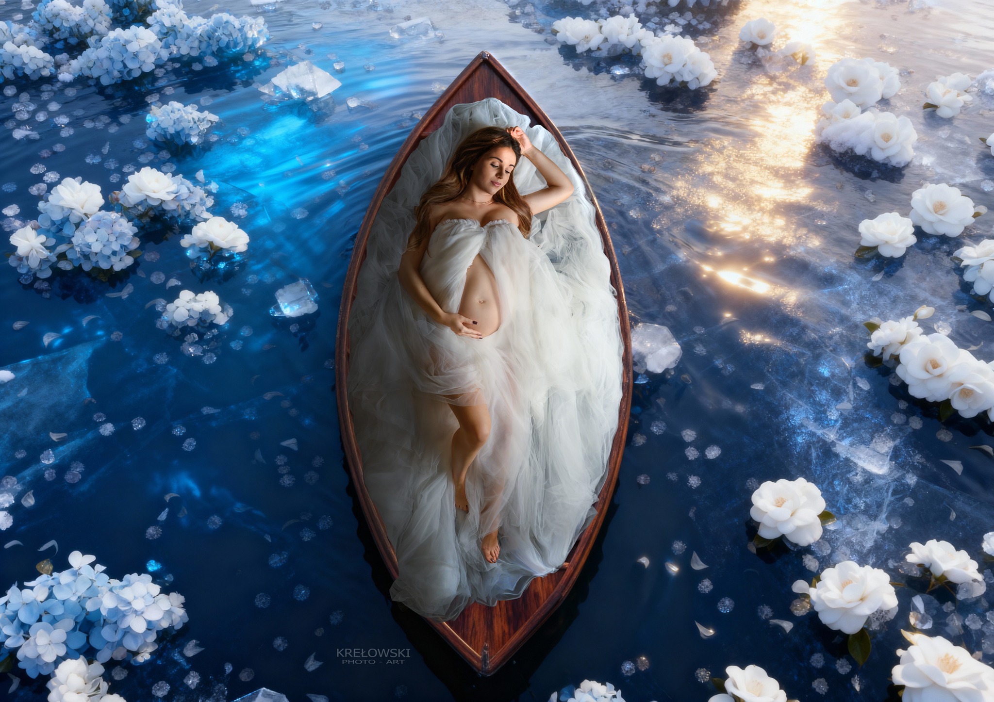 Kobieta w ciąży, otulona białym materiałem, leży w łodzi na wodzie otoczonej białymi kwiatami i kryształkami lodu. Ujęcie z góry, miękkie światło.
