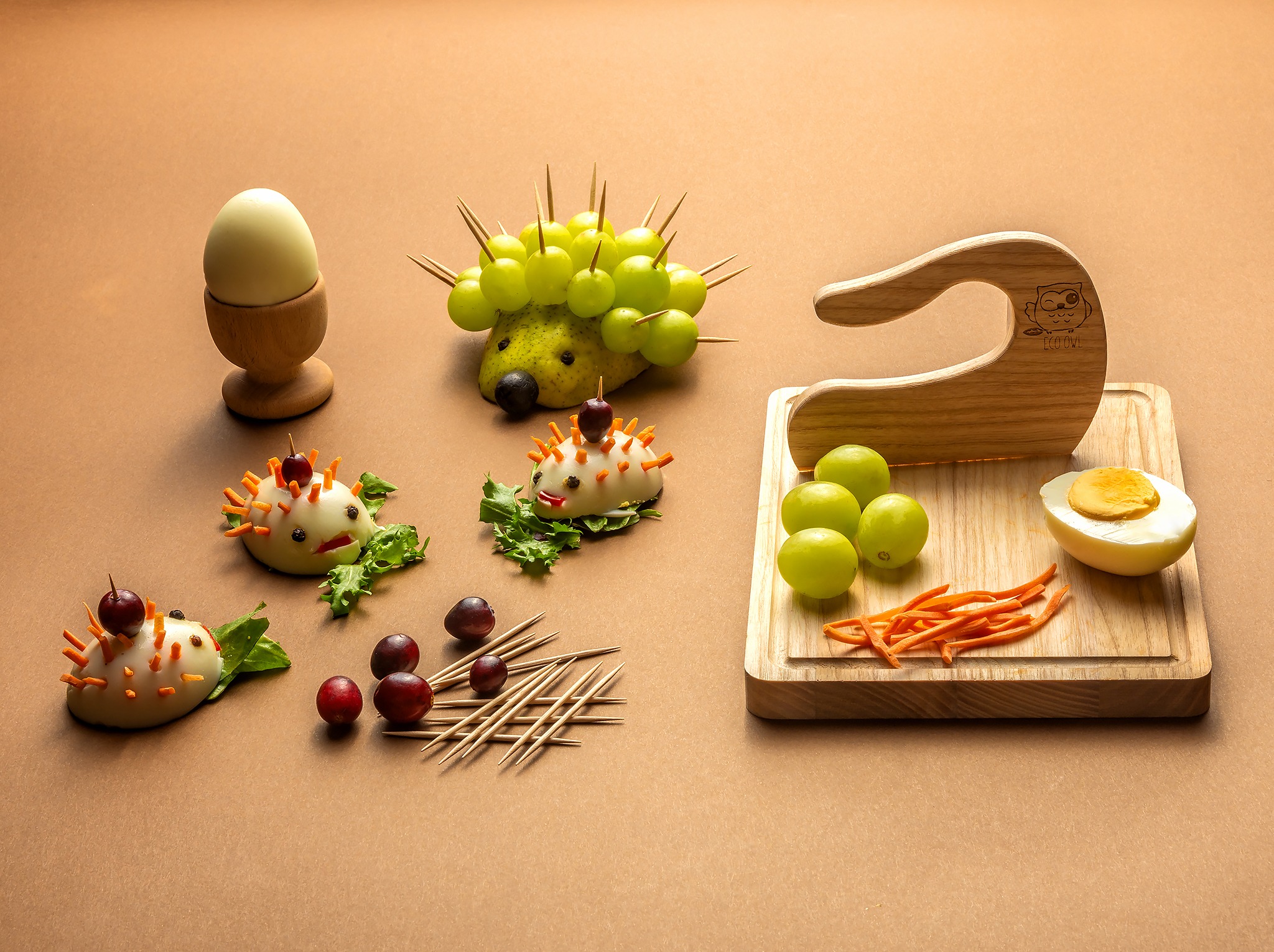 Kreatywna kompozycja z jajek i owoców uformowanych w kształt zwierząt, na brązowym tle. Jajko w kieliszku, jeż z gruszek i winogron, jeże z jajek i marchewki, drewniana deska z jajkiem.