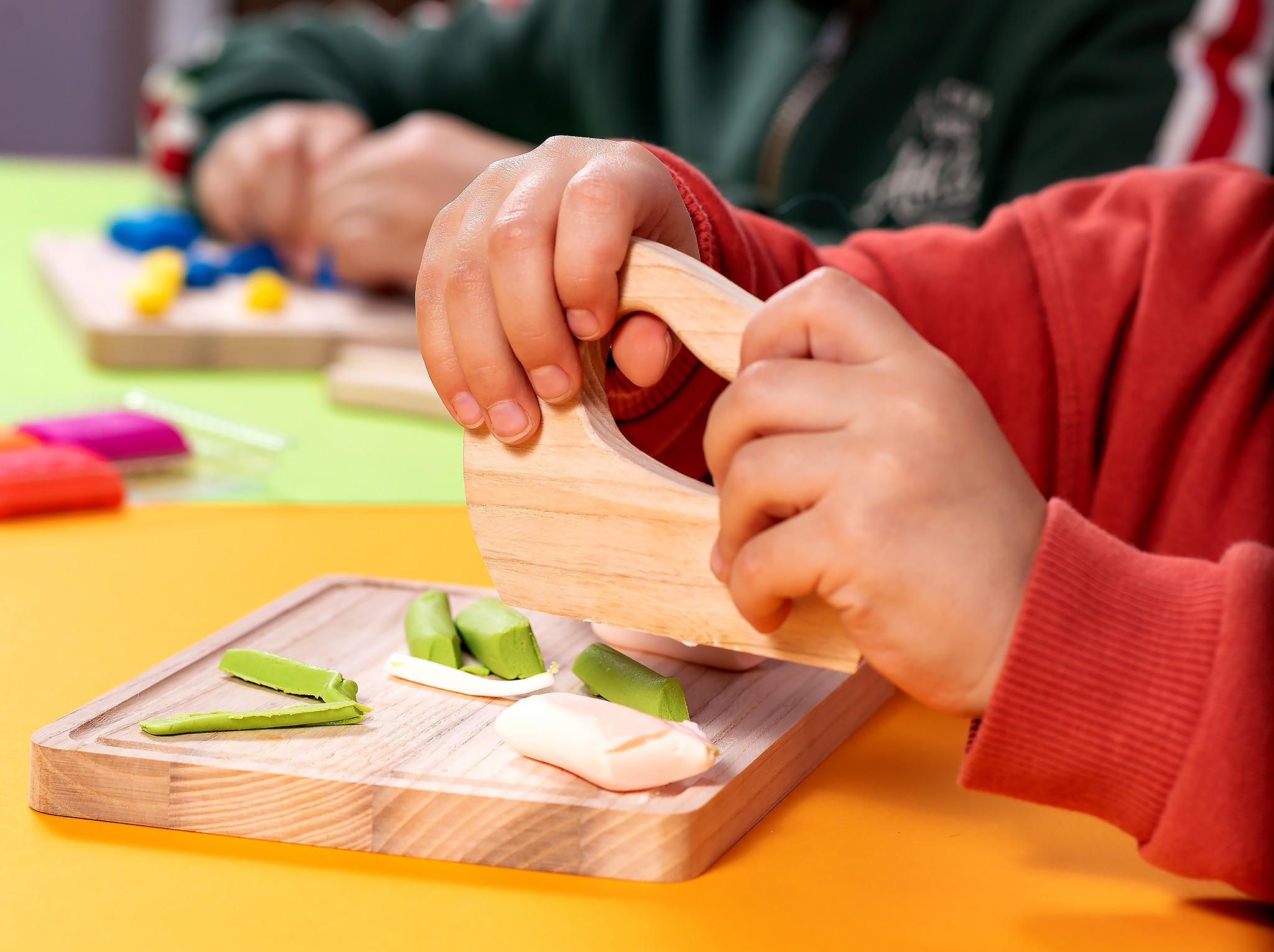 Dziecko w czerwonej bluzie formuje kolorową plastelinę na drewnianej desce za pomocą drewnianego narzędzia. W tle inny uczestnik zajęć.