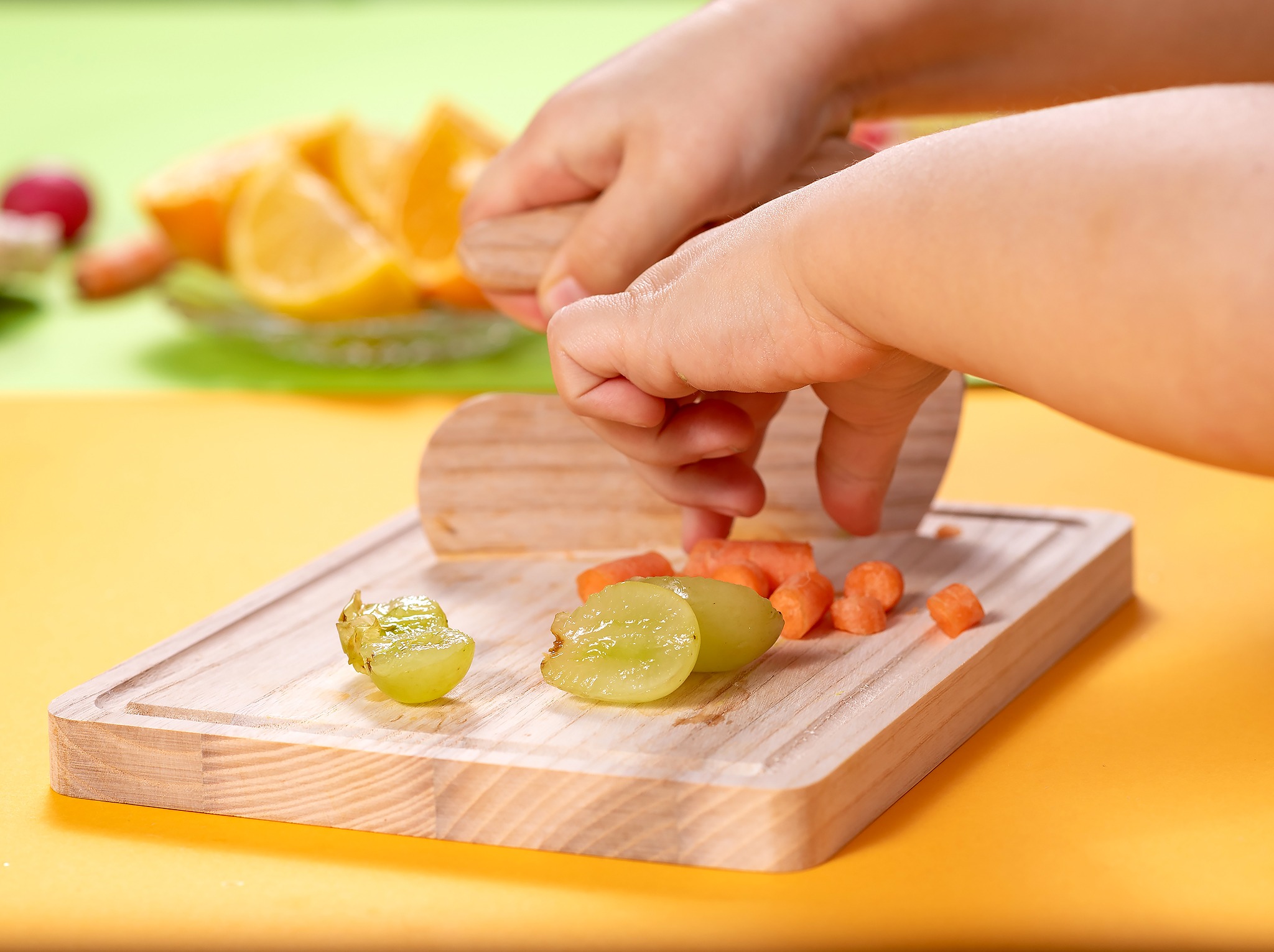Dziecięce dłonie kroją drewnianym nożem winogrona i marchewkę na desce. W tle pomarańcze na talerzu, na żółto-zielonym tle.