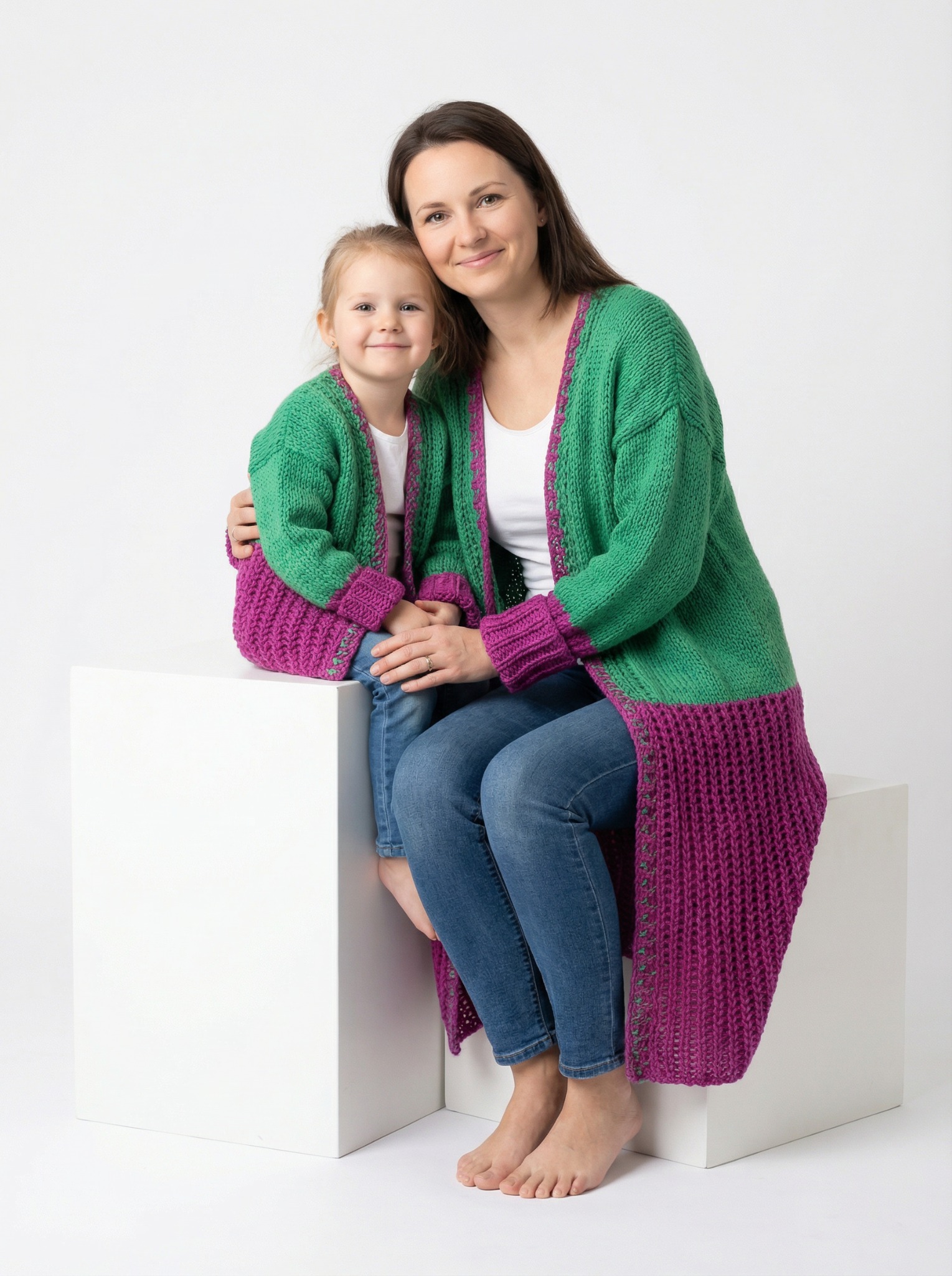 Uśmiechnięta mama obejmuje córkę, obie w dopasowanych, dzierganych, zielono-fioletowych kardiganach na białym tle. Sesja w jasnym, studyjnym oświetleniu.