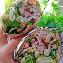 Red Target sp. z o o - Dwa tortille wrap z kurczakiem, bekonem i świeżymi warzywami, trzymane w dłoni na tle zielonej rośliny. Jeden wrap owinięty folią aluminiową.