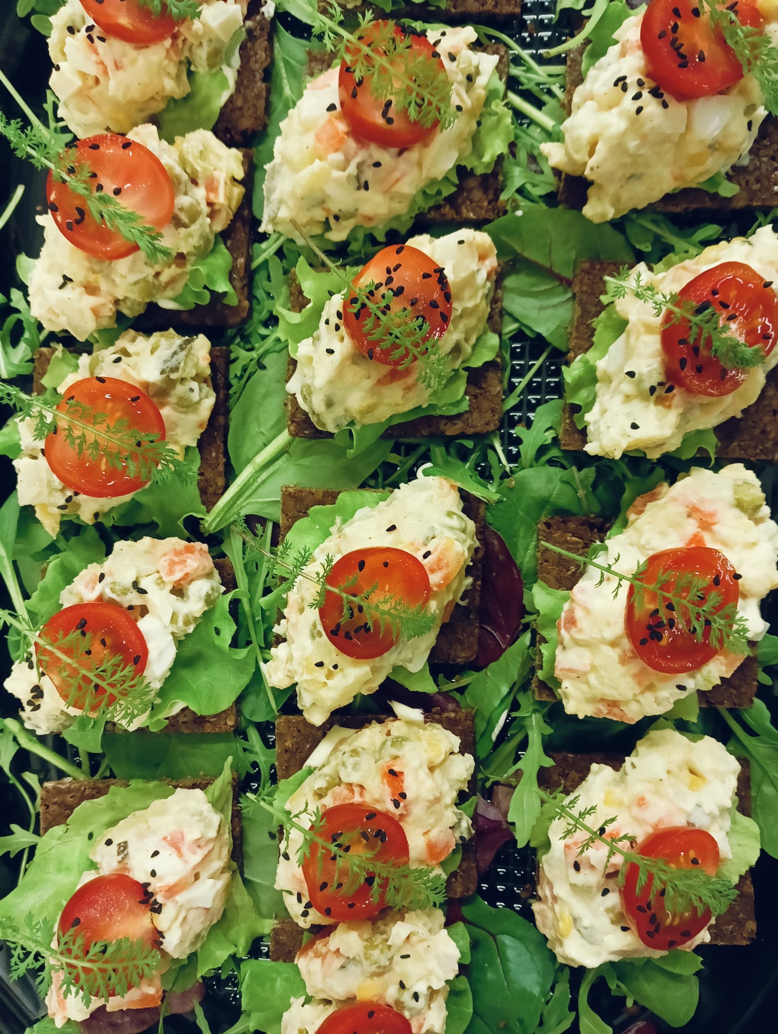 Apetyczne kanapeczki na ciemnym pieczywie z sałatką jarzynową, pomidorkiem cherry i koperkiem, ułożone na świeżych liściach sałaty. Elegancka kompozycja kulinarna.