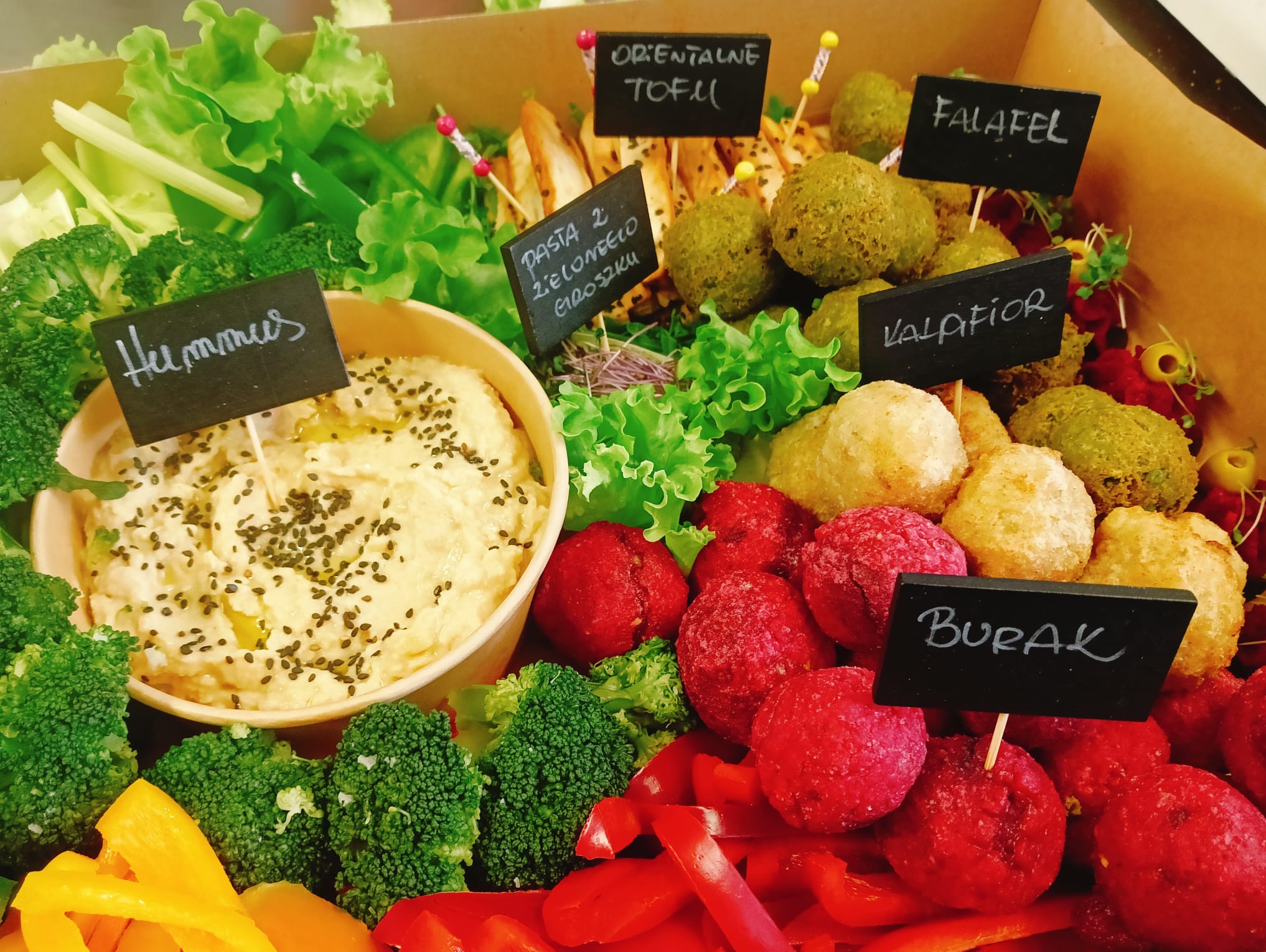 Kolorowa kompozycja wegańskich przekąsek w pudełku: hummus, falafel, tofu orientalne, pasta z zielonego groszku, buraki, brokuły i papryka. Etykiety z nazwami.