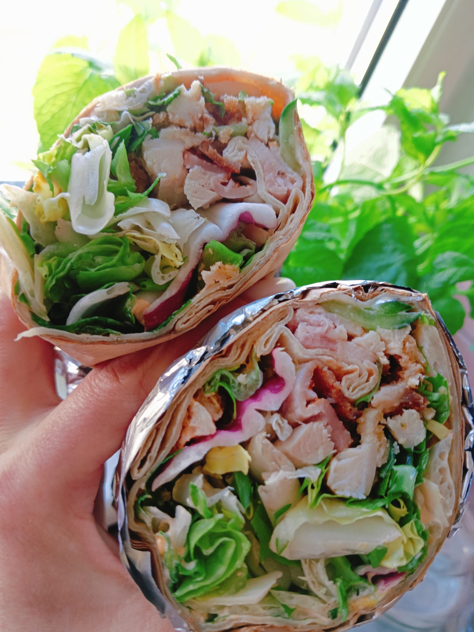 Dwa tortille wrap z kurczakiem, bekonem i świeżymi warzywami, trzymane w dłoni na tle zielonej rośliny. Jeden wrap owinięty folią aluminiową.