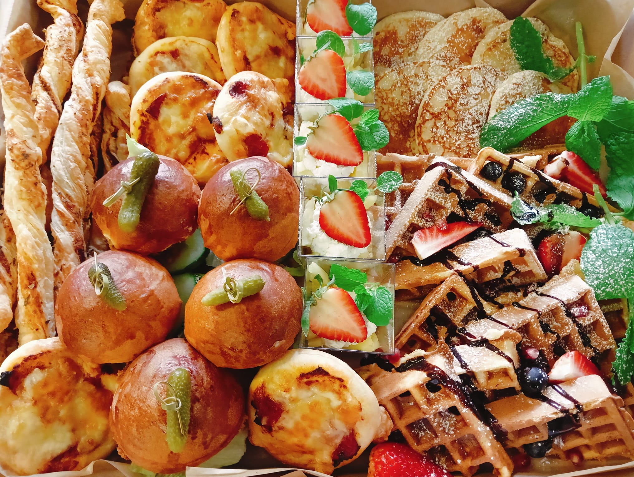 Bogaty wybór przekąsek w pudełku: mini burgery z piklami, tartaletki, naleśniki, gofry z owocami i desery w kubeczkach, udekorowane miętą. Idealne na catering.