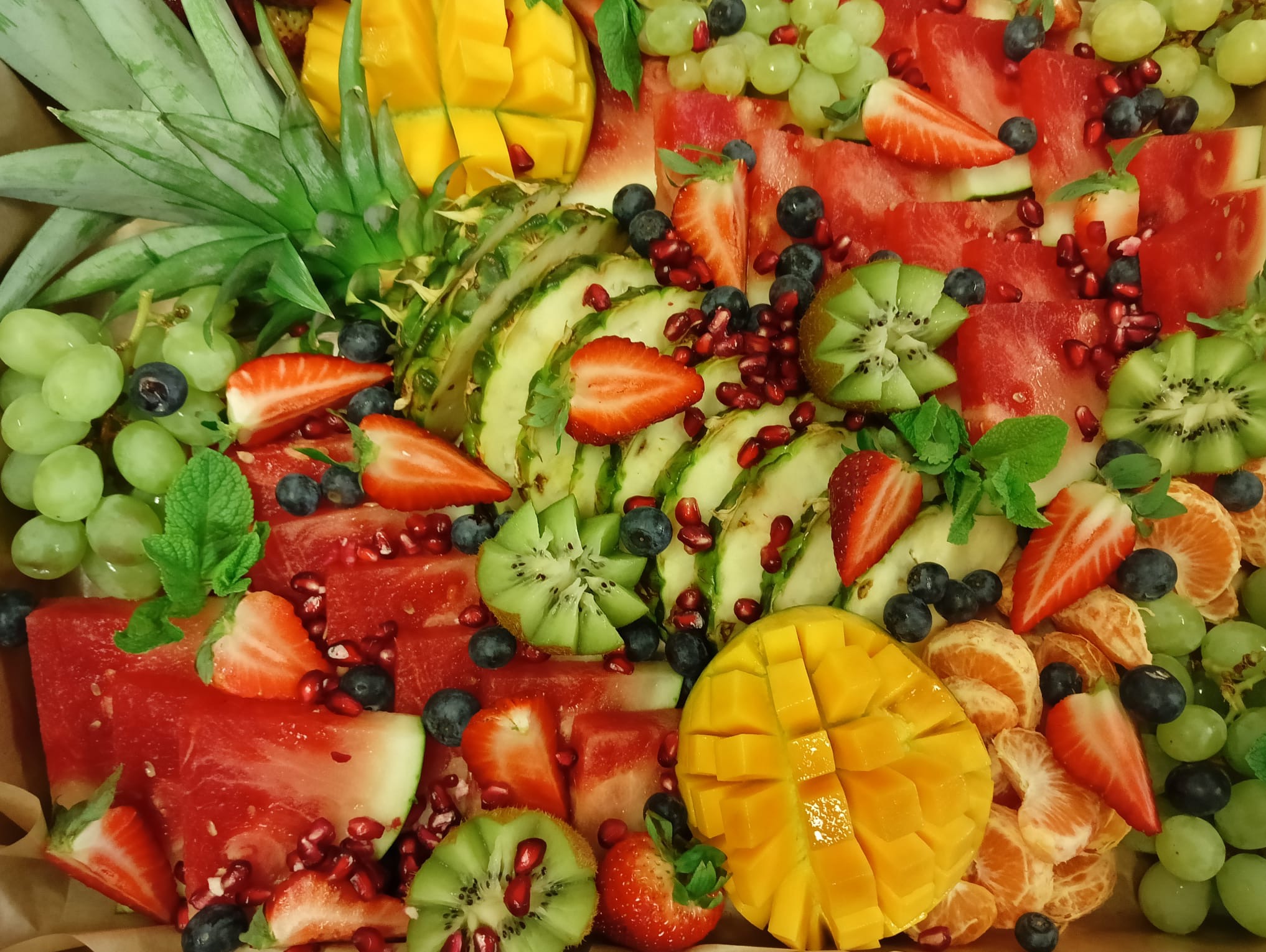 Bogata kompozycja owocowa: mango w kostkach, arbuz, kiwi, truskawki, winogrona i borówki, udekorowana granatem i miętą. Idealna na catering lub słodki stół.