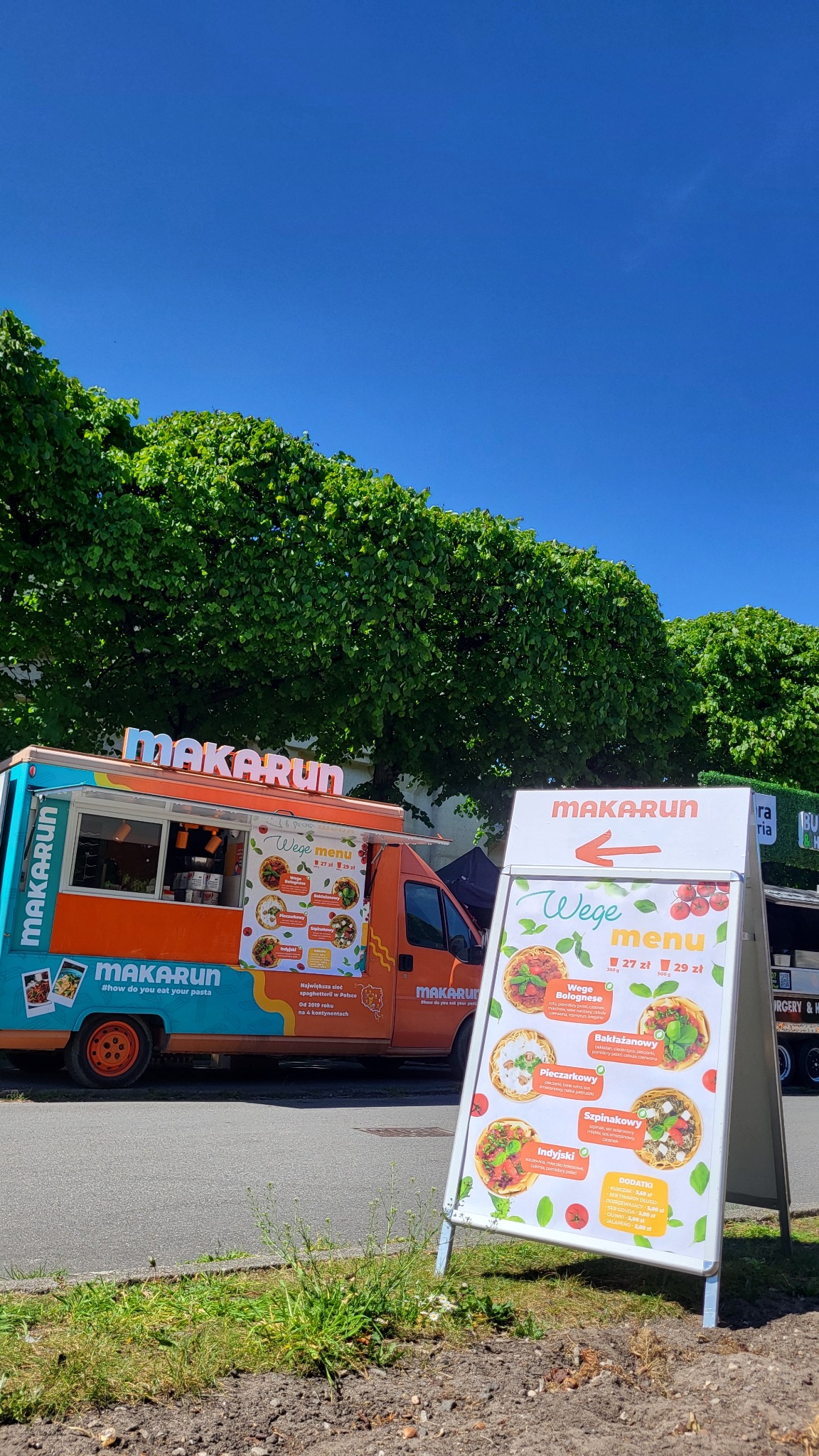 Food truck Makarun z wege menu we Wrocławiu. Kolorowy pojazd i tablica z ofertą dań wegetariańskich na tle zieleni i błękitnego nieba.