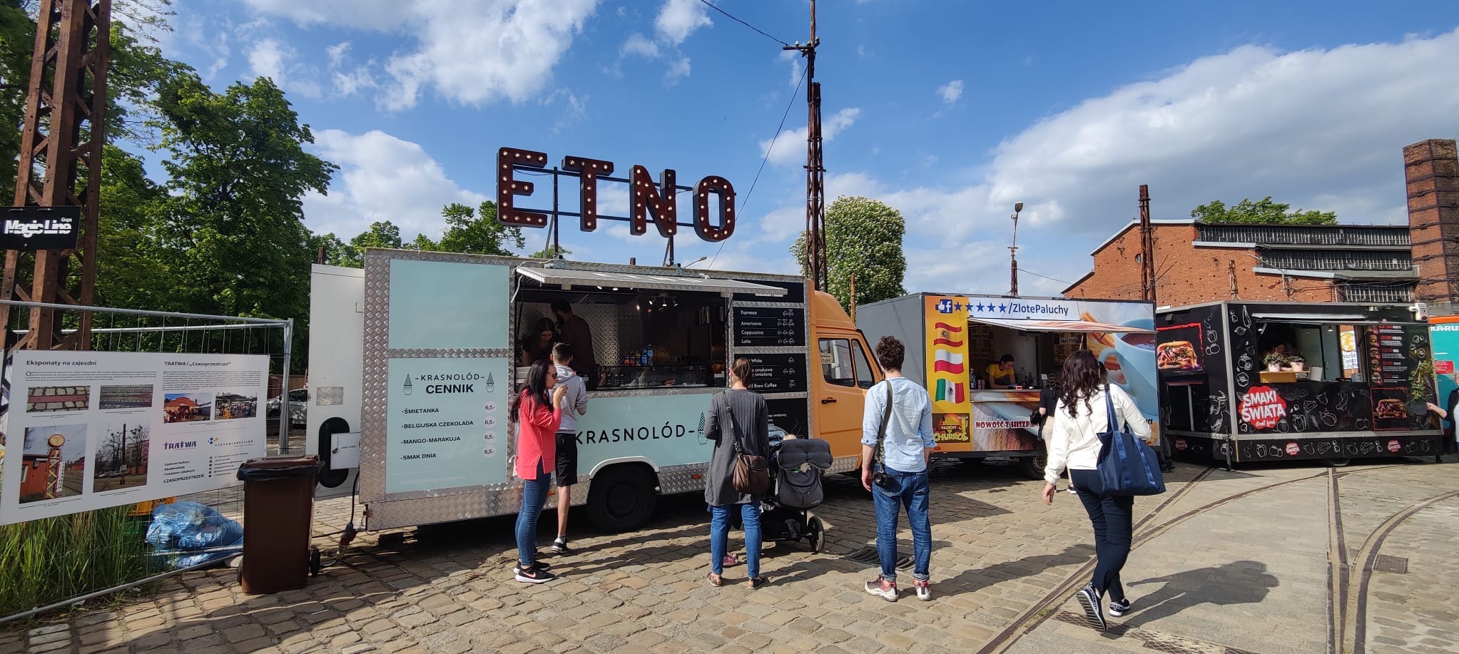 Food trucki z ofertą gastronomiczną na zewnątrz, w tle ceglany budynek. Na pierwszym planie klienci przed food truckiem 'ETNO' z menu lodów.