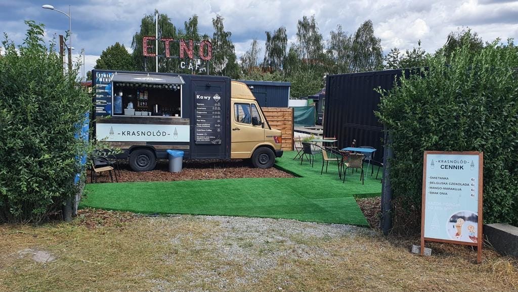 Mobilna kawiarnia 'Krasnolód' w food trucku z menu kaw i cennikiem lodów, otoczona zielenią i stolikami na zewnątrz. Letnia atmosfera.