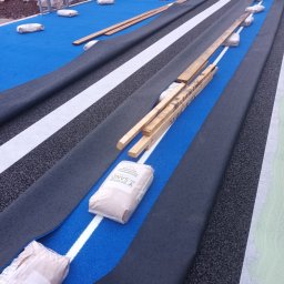 Usługi Monterskie i Budowlane Bartosz Świerszczewski - Montaż nawierzchni sportowej: rozłożone pasy czarnej gumy, białe linie, worki z piaskiem i drewniane listwy na niebieskim podłożu. Etap prac wykończeniowych.