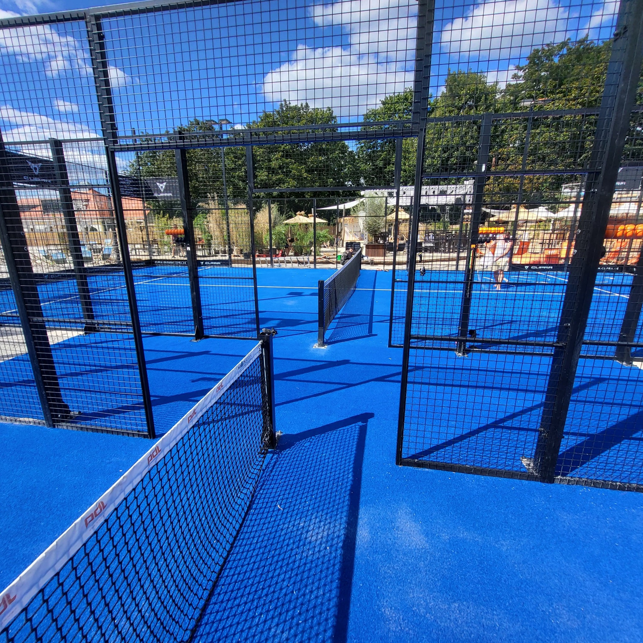 Niebieski kort do padla z czarną siatką i ogrodzeniem, widok z perspektywy gracza, w tle zieleń i budynki pod błękitnym niebem.