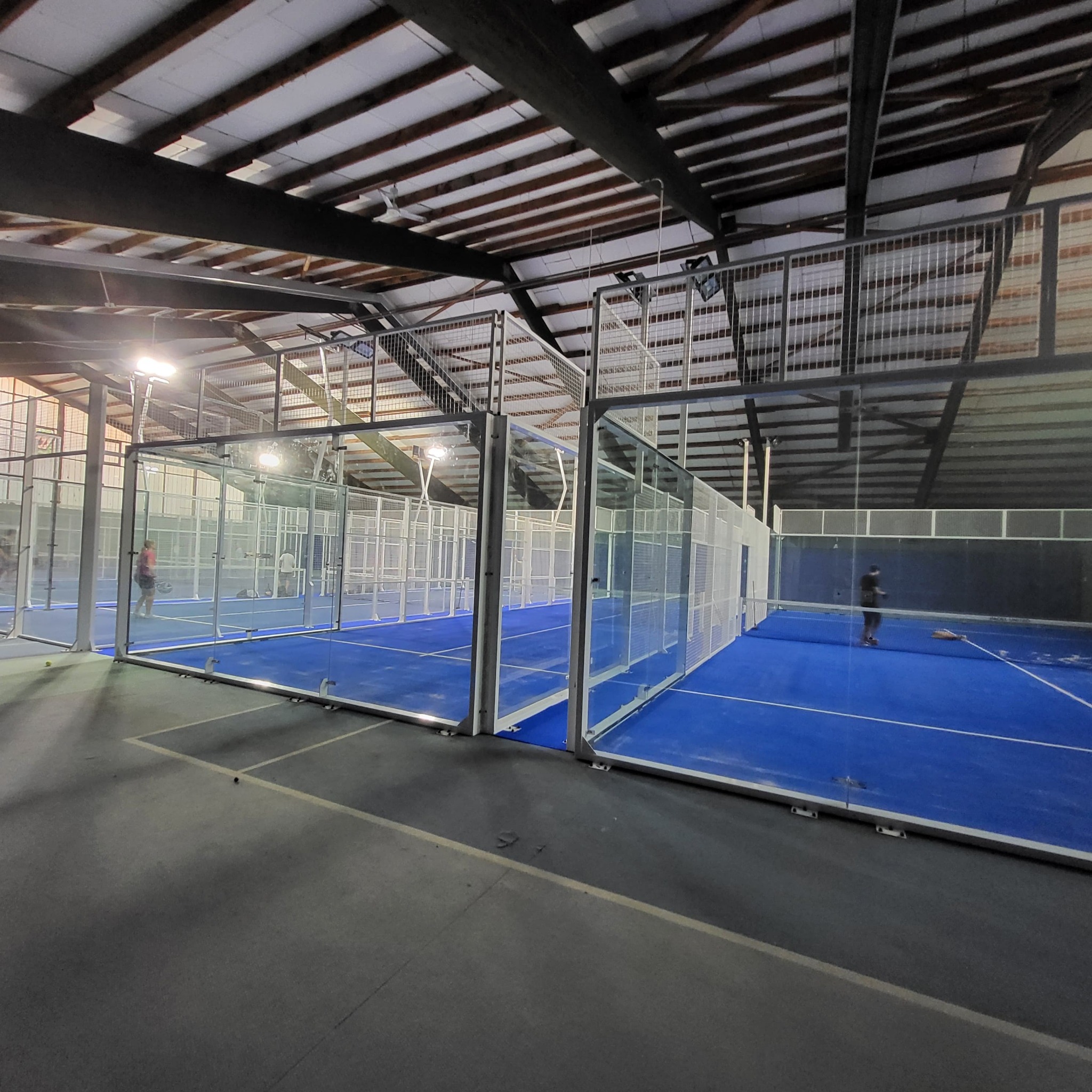 Hala sportowa z kortami do padla, oświetlona lampami. Widoczne szklane ściany kortów i niebieska nawierzchnia. Konstrukcja dachu z belek stalowych i drewnianych.