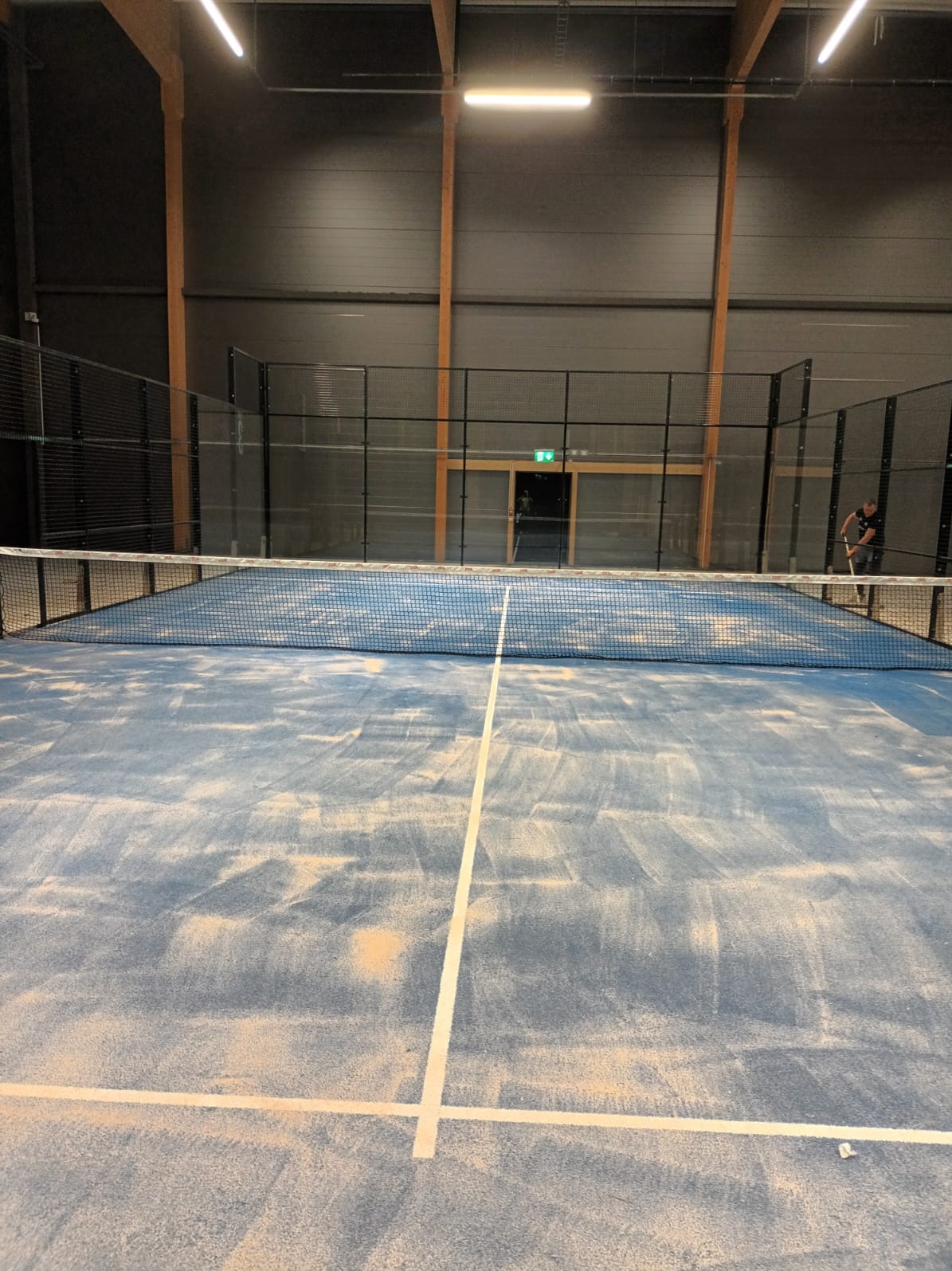 Wnętrze hali sportowej z kortem do padla, siatką i widocznymi liniami. W tle osoba sprzątająca kort. Oświetlenie górne, drewniane elementy konstrukcyjne.
