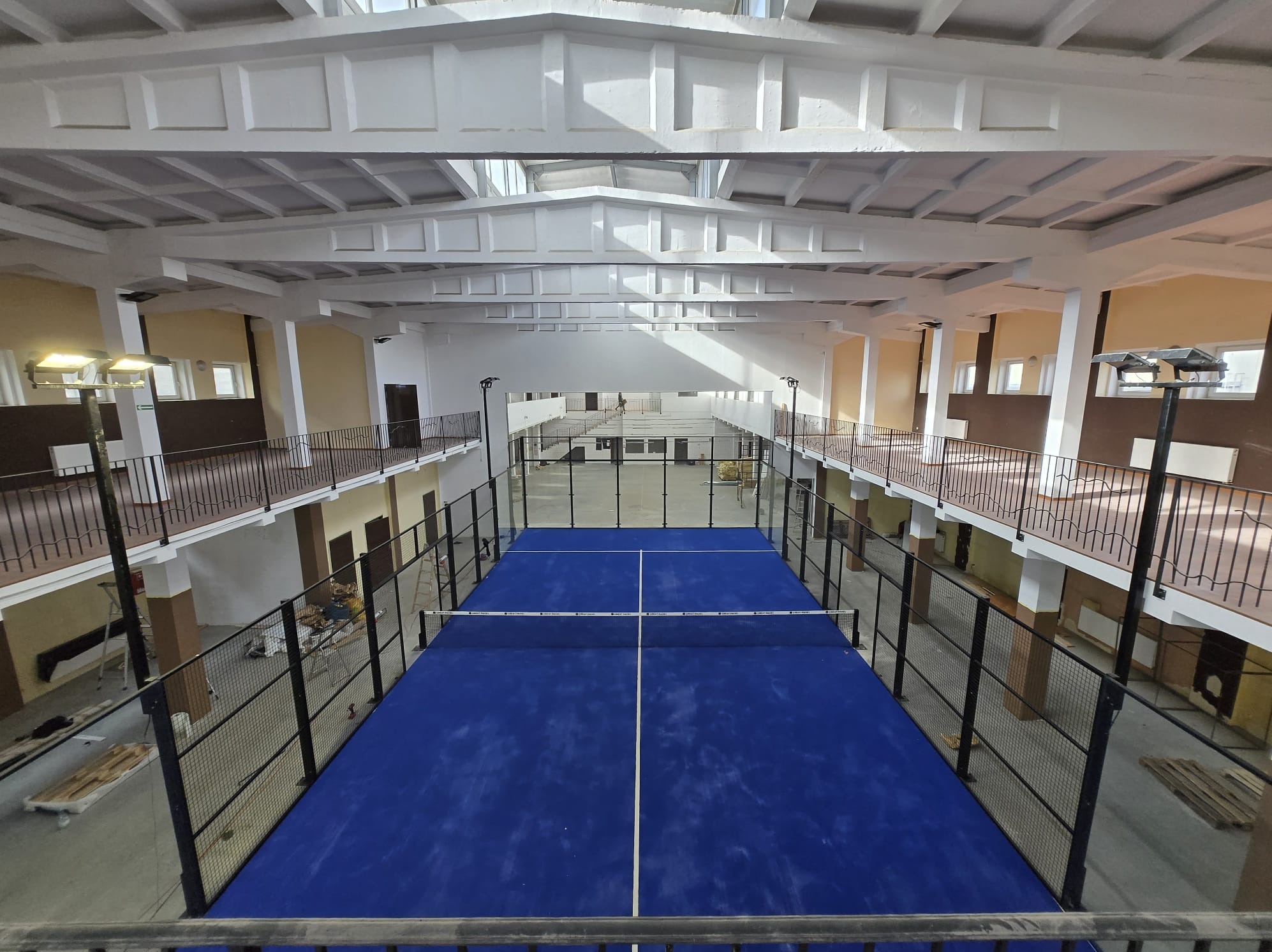 Widok z góry na niebieski kort do padla w przestronnym, jasnym wnętrzu hali sportowej z balkonami i białymi kolumnami. Siatka na korcie.