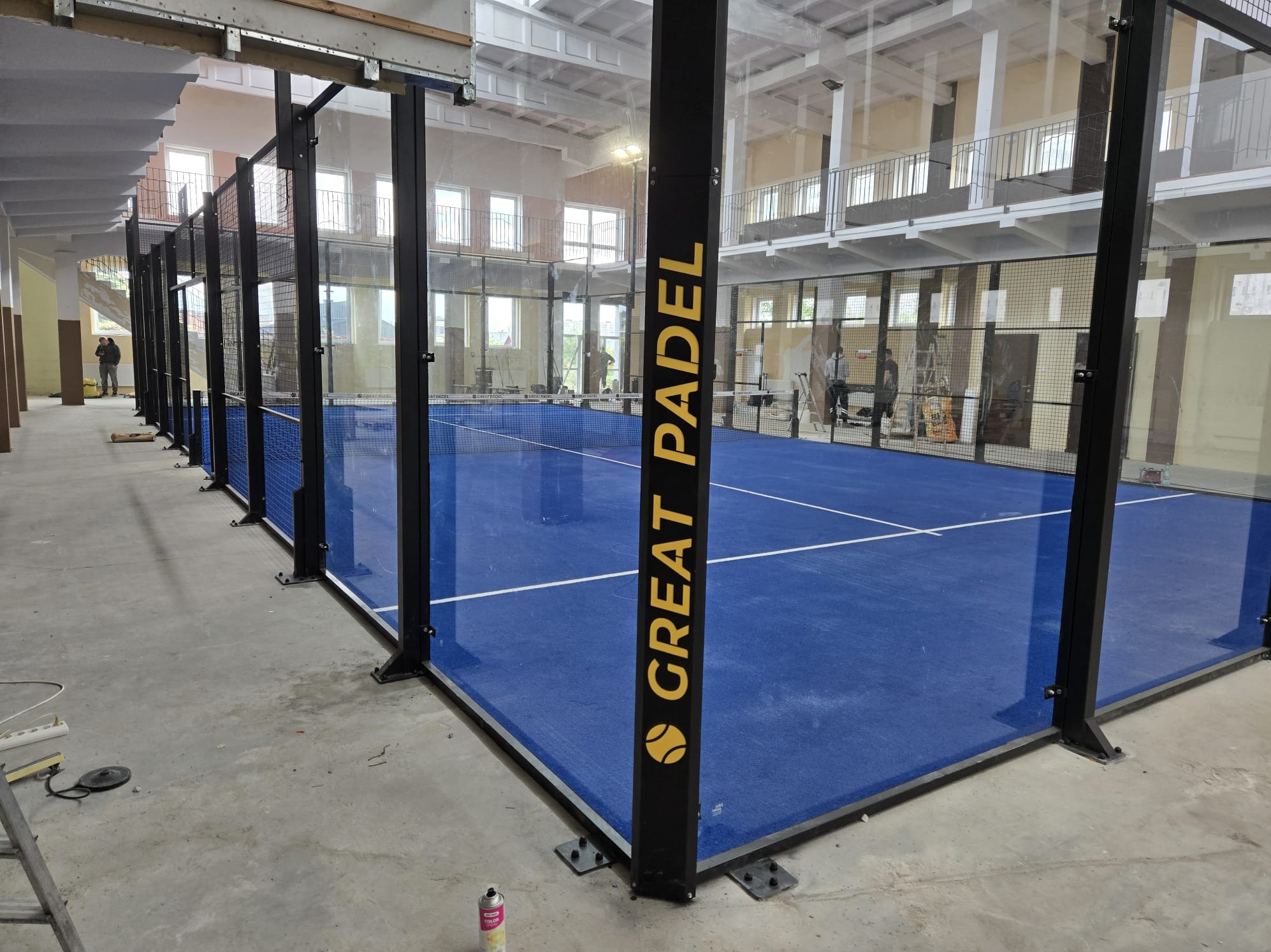 Nowoczesne boisko do padla z niebieską nawierzchnią, otoczone szklanymi ścianami i czarną metalową konstrukcją. Widoczne elementy montażowe i wykończeniowe.