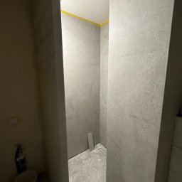 ROB-BUD Robert Kaczyński - Wykończona w szarościach kabina prysznicowa z odpływem liniowym, zabezpieczona żółtą taśmą malarską przy suficie, w trakcie prac wykończeniowych w łazience.