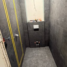 ROB-BUD Robert Kaczyński - Ciemna łazienka w trakcie remontu: szare płytki, żółte taśmy wyznaczające obszar, przygotowane instalacje pod WC i umywalkę. Widoczne narzędzia i notatki na ścianie.